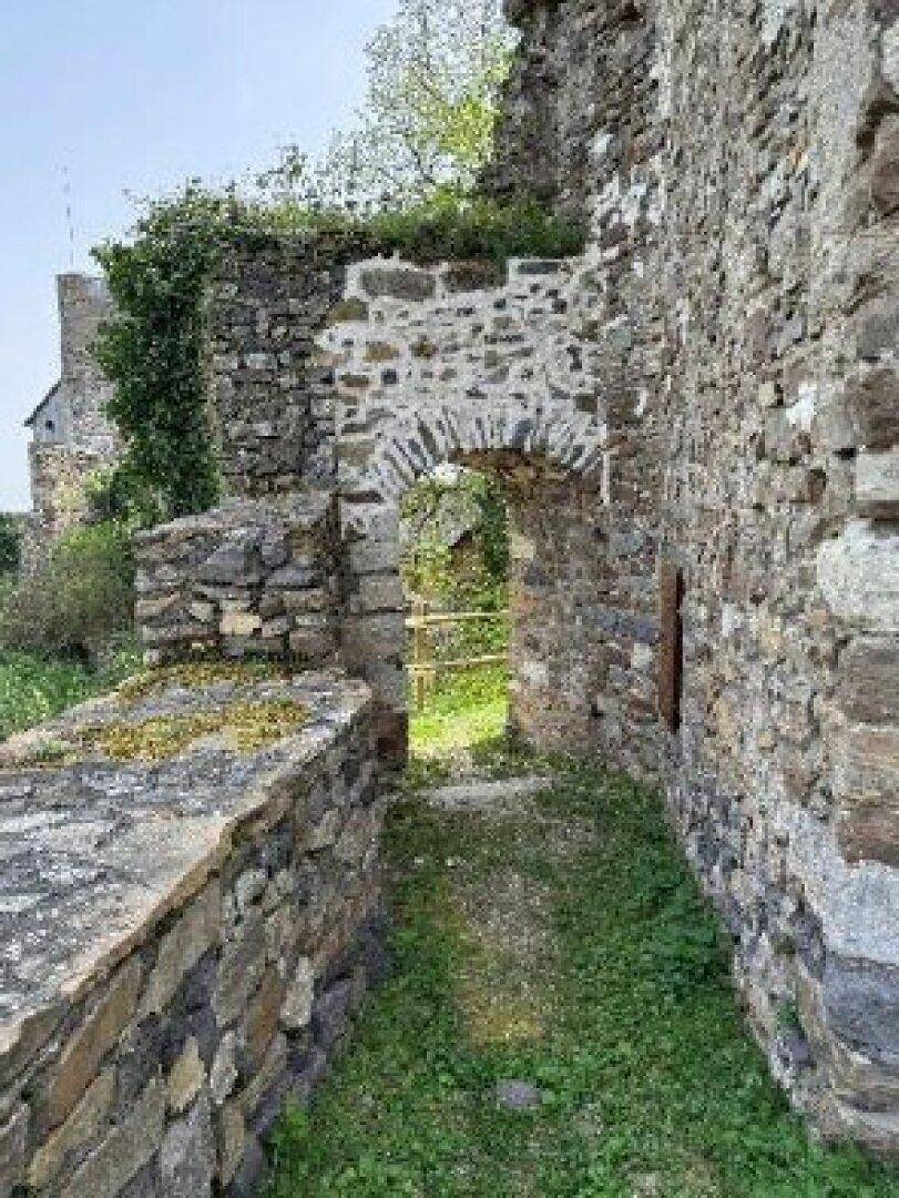Ein schmaler Weg entlang alter Steinmauern einer Burgruine mit einem weiteren Torbogen in der Ferne.
