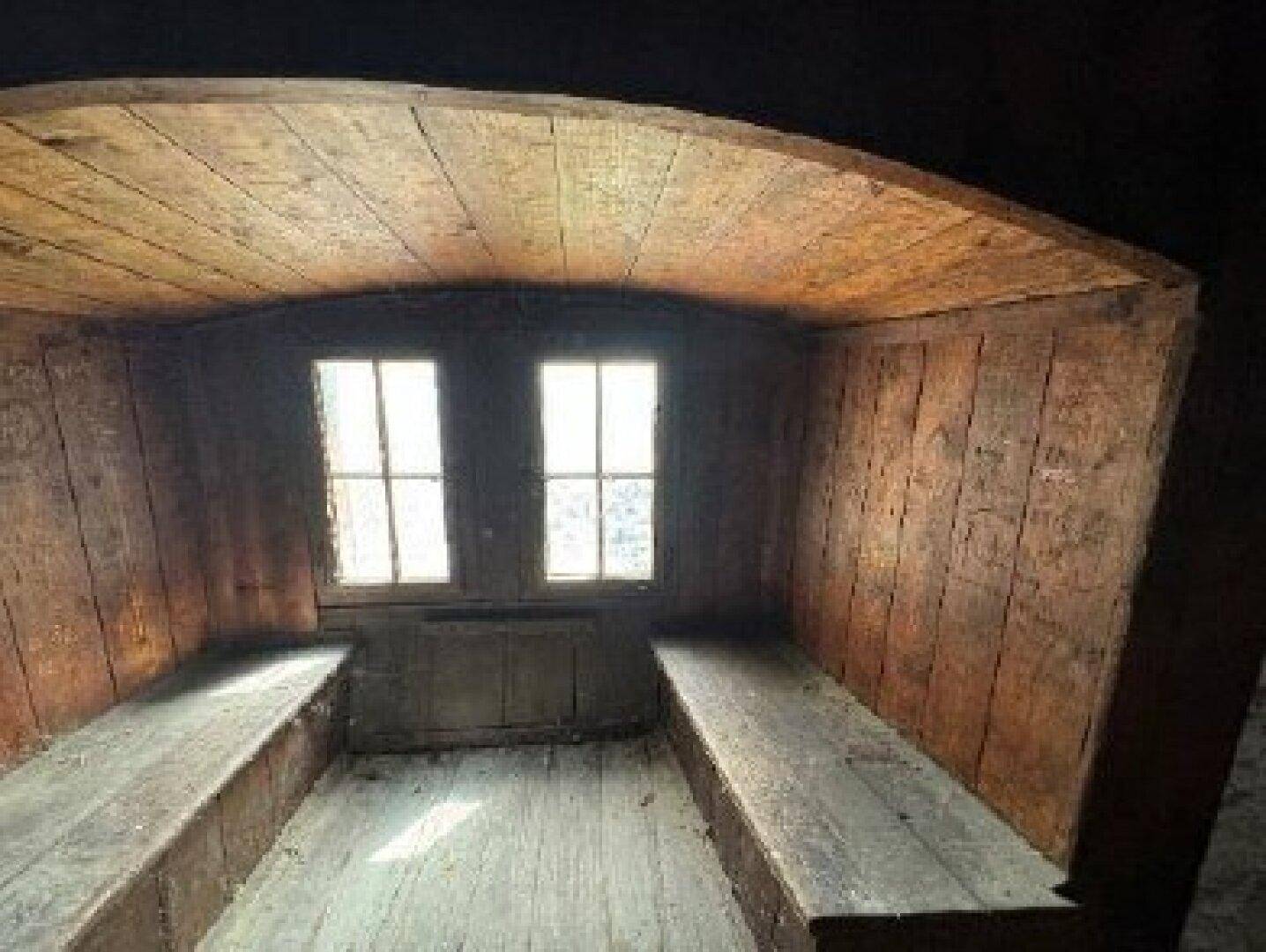 Kleiner, rustikaler Raum mit Holzwänden, Holzbänken und zwei Fenstern, die Licht hereinlassen.