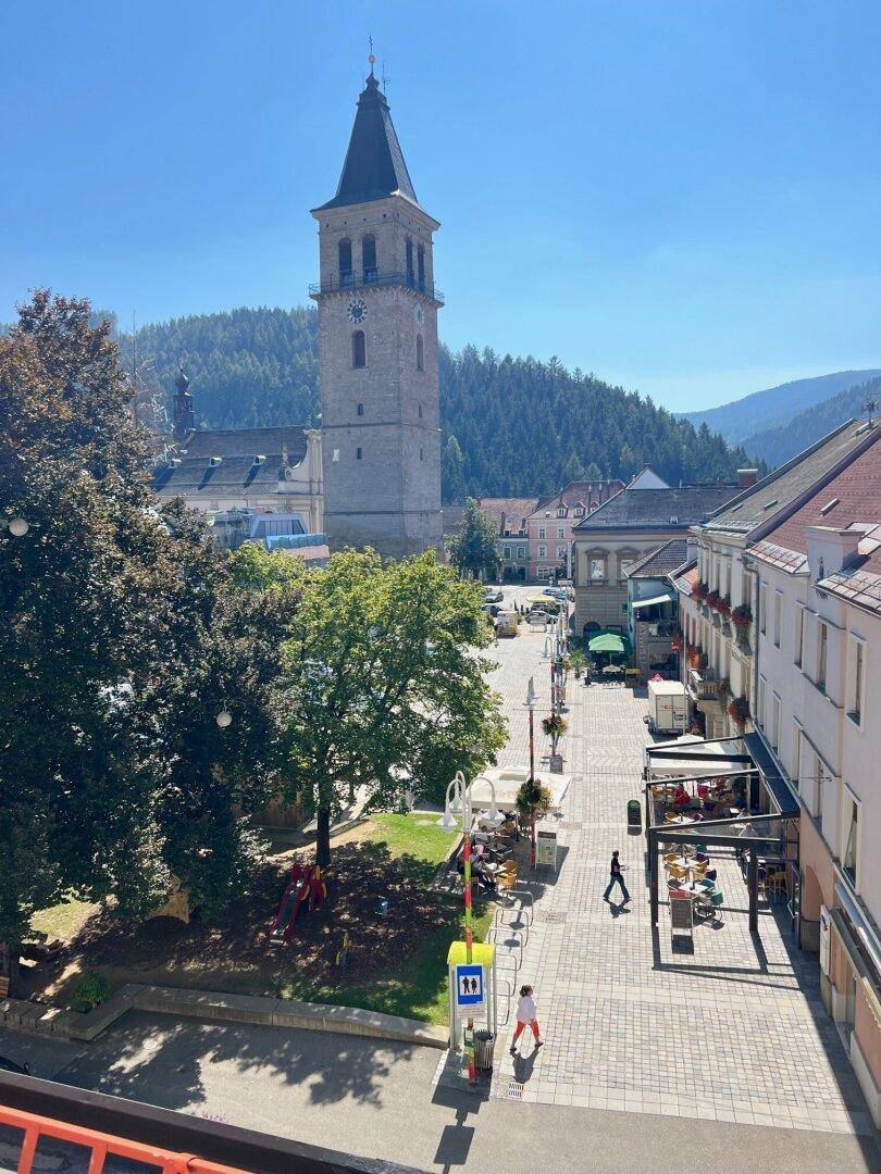 Helle Tagesansicht des Stadtplatzes mit historischem Kirchturm, Bergen und belebter Fußgängerzone.