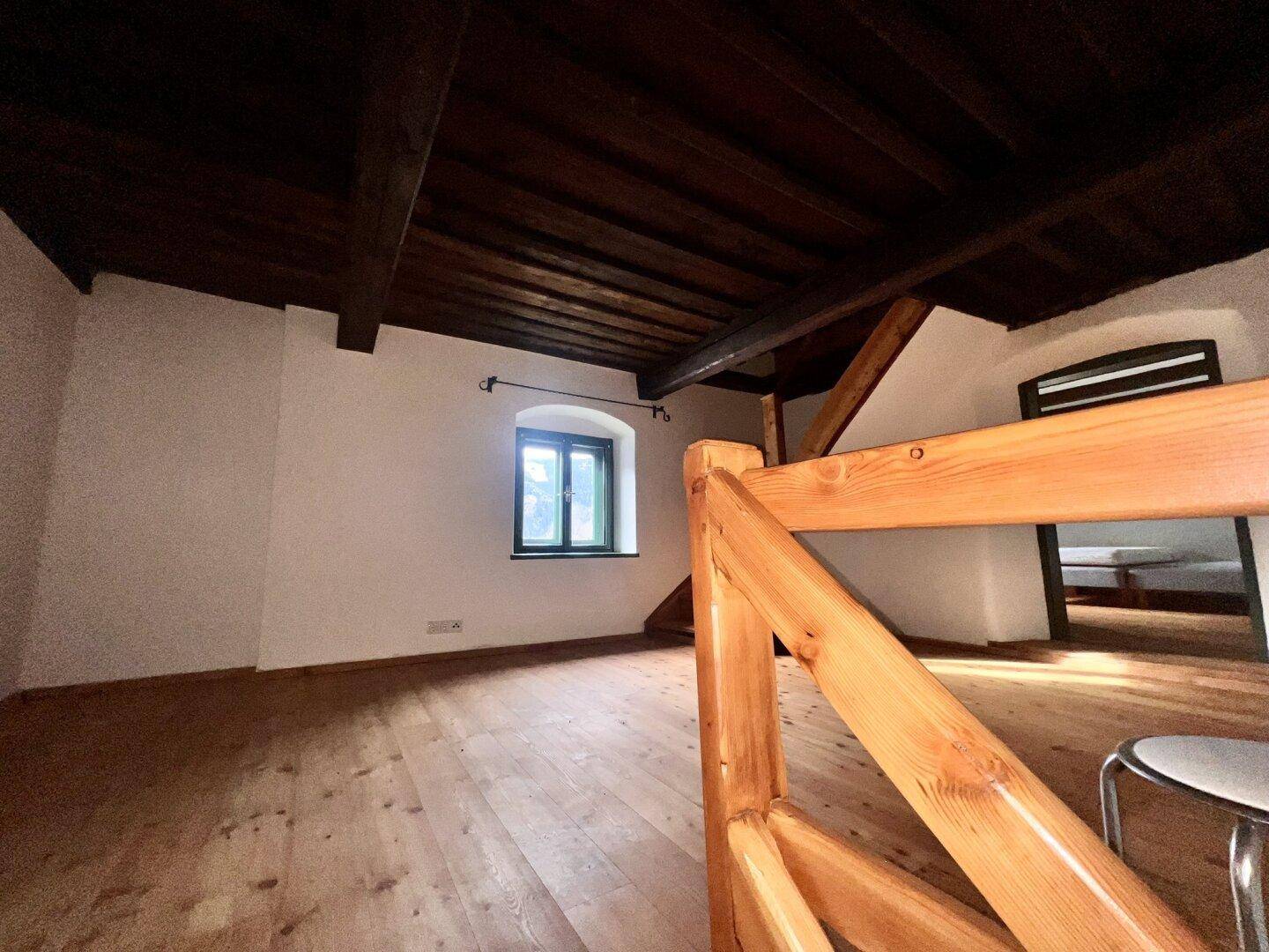 Offener Bereich im Obergeschoss mit Holzdielenboden, sichtbaren Balken und Blick auf ein Fenster.
