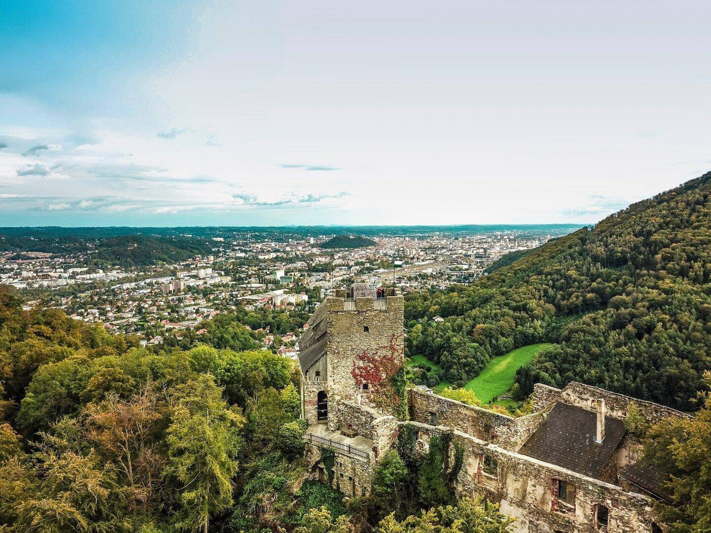 Panoramablick auf eine alte Burgruine mit einer Stadt und weiten Berglandschaft im Hintergrund.