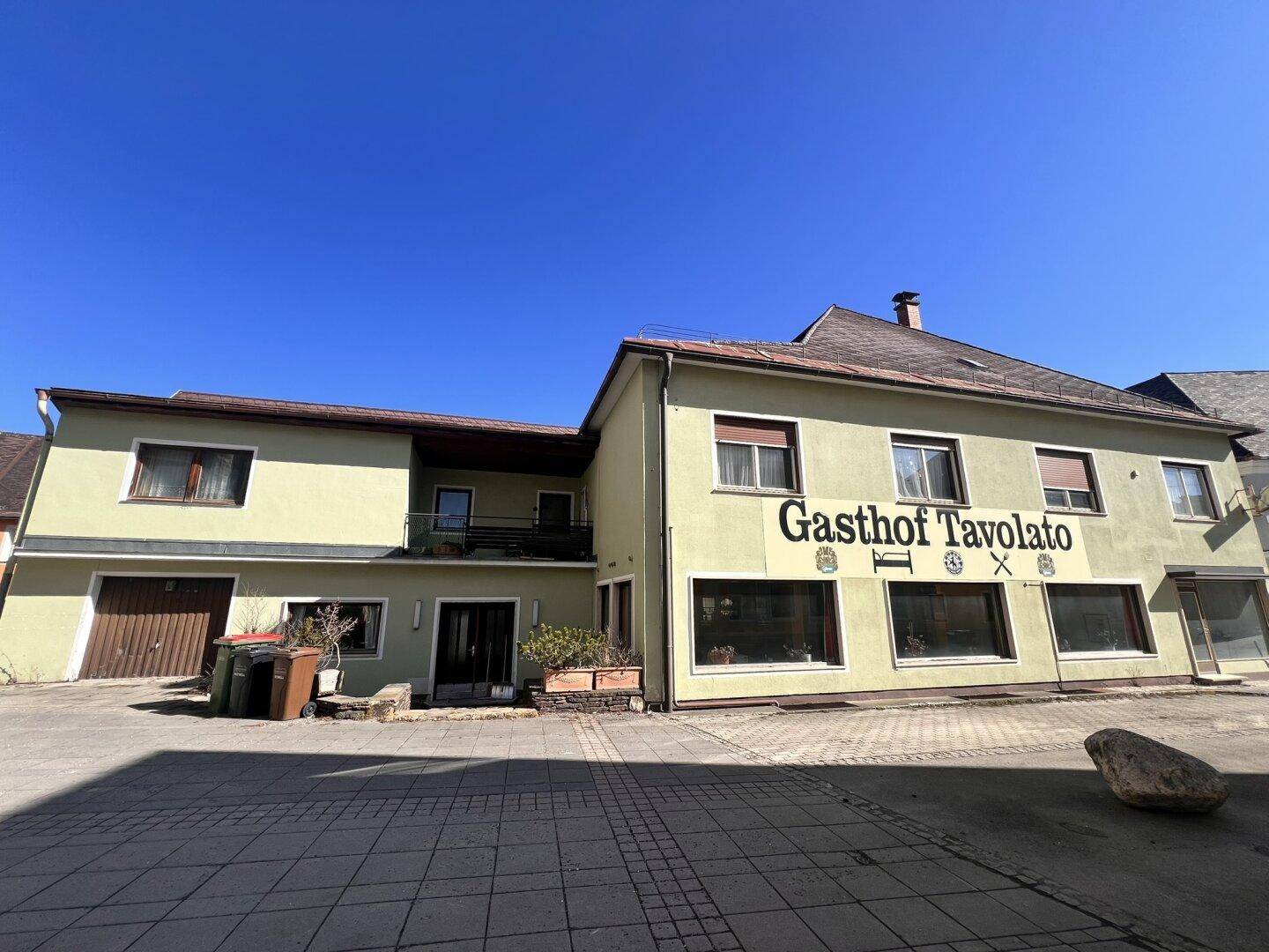 Rückseite des Gasthofs mit Garagentor und gepflastertem Bereich an einem sonnigen Tag.