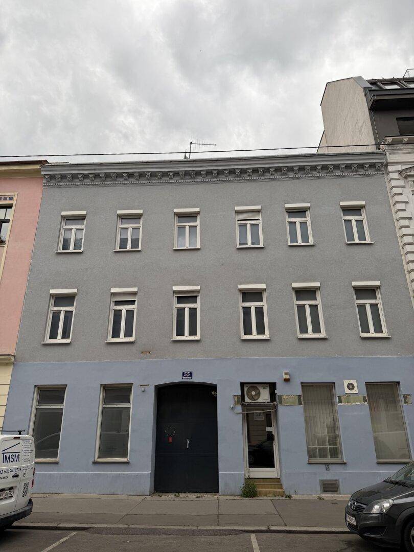 Nahaufnahme einer grauen Gebäudefassade mit blauer Sockelzone, weißen Fenstern und einer dunklen Eingangstür.