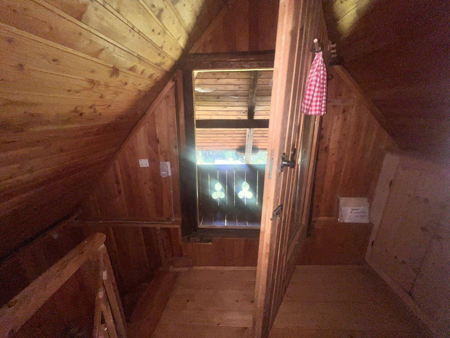 Rustikaler Zugang zum Dachboden mit Holztäfelung und Blick durch eine kleine Öffnung nach draußen.