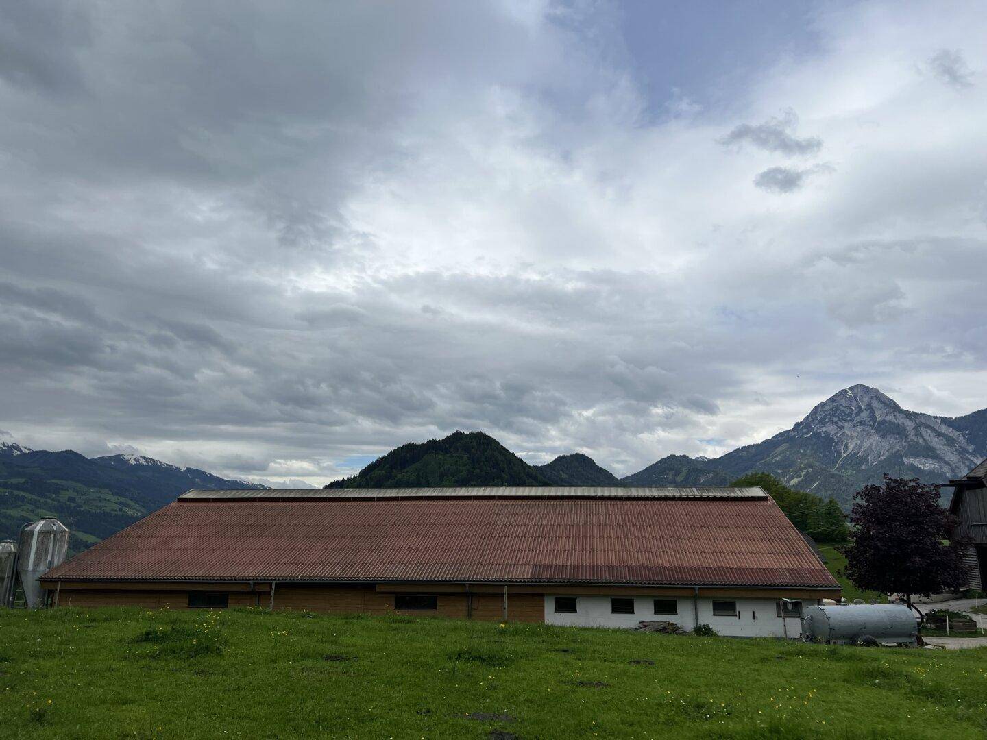 Langes landwirtschaftliches Gebäude mit rotem Ziegeldach, umgeben von grünen Wiesen und majestätischen Bergen.