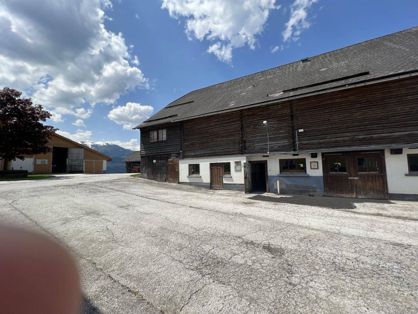 Großes Holzgebäude mit traditioneller Fassade und weitem Vorplatz, umgeben von ländlicher Berglandschaft.