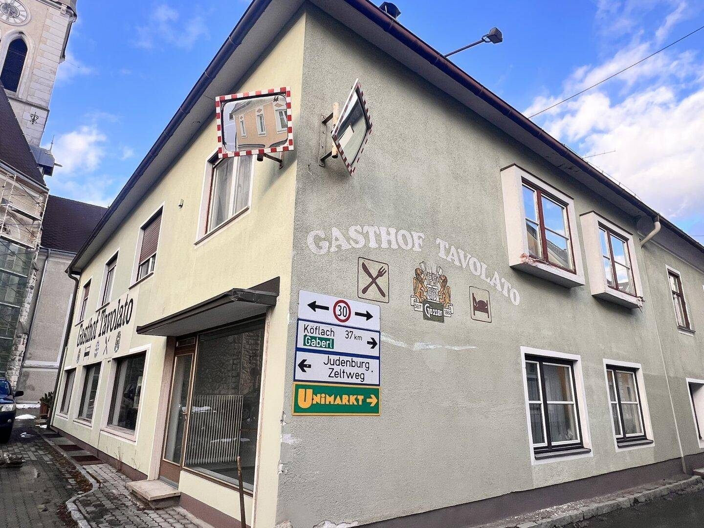 Seitenansicht des Gasthofs mit Wegweisern und einem Verkehrsspiegel an der Ecke.