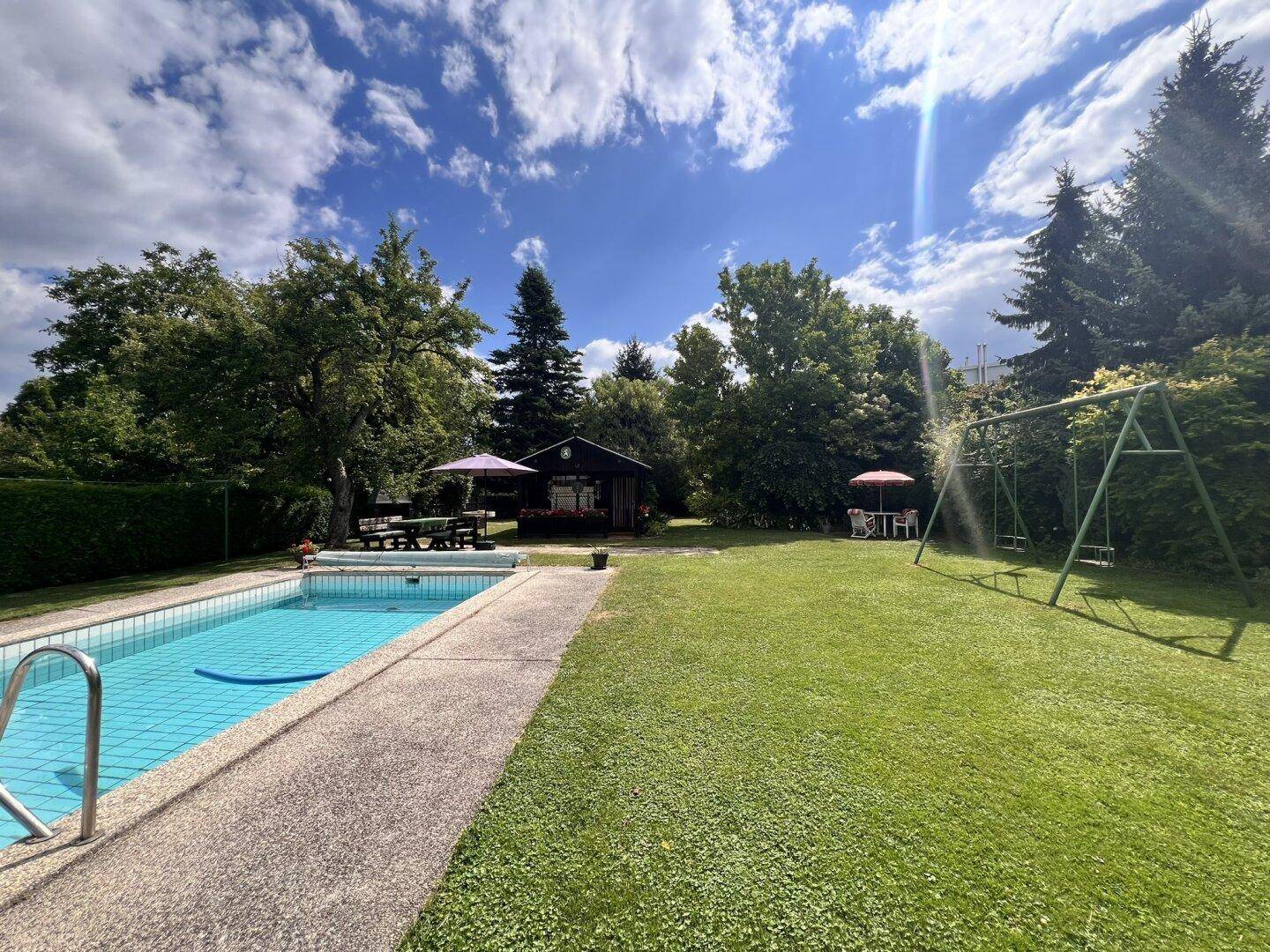 Weitläufiger Garten mit Pool, Gartenhaus und altem Baumbestand unter blauem Himmel.