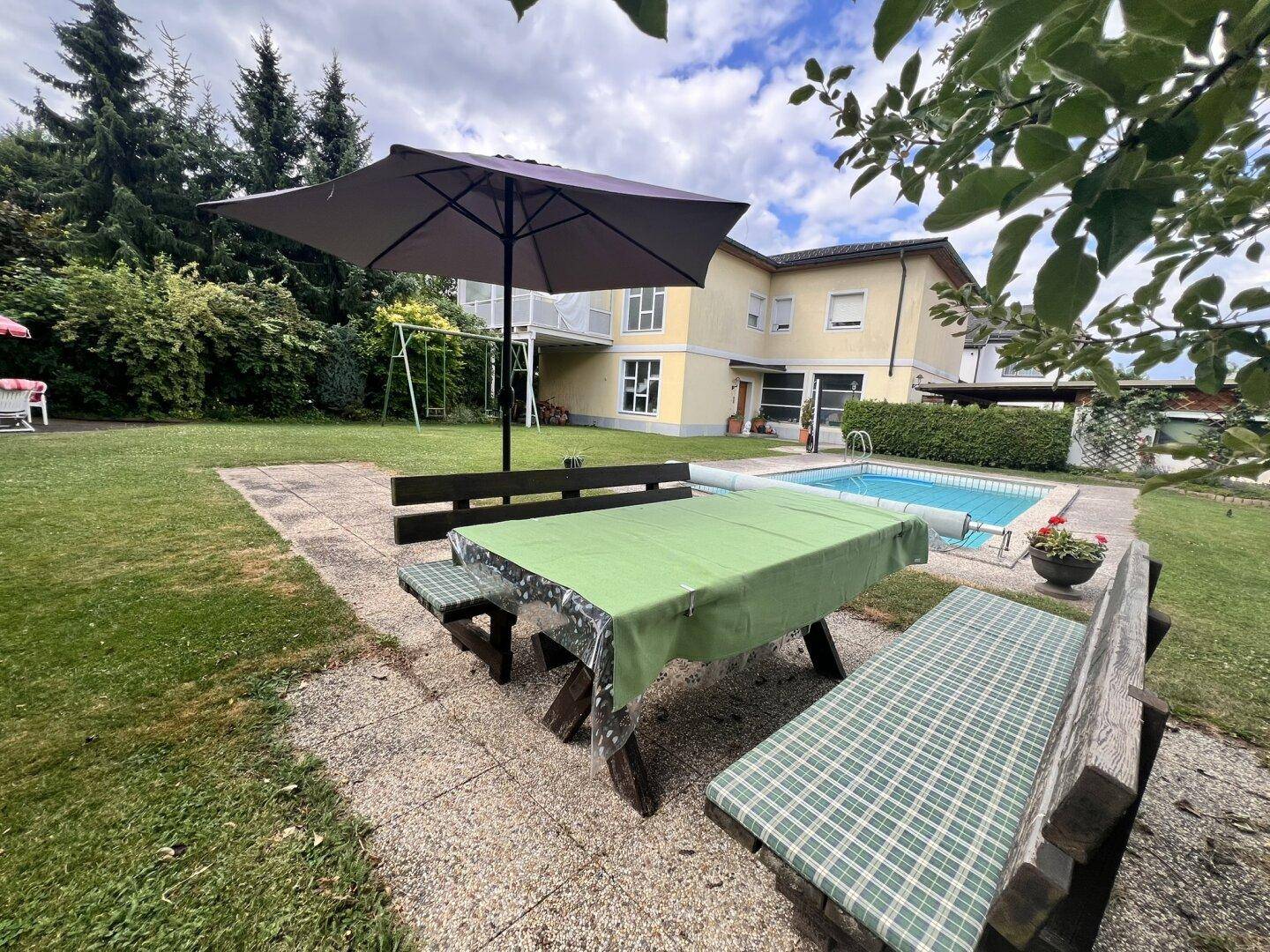 Gemütlicher Essbereich im Freien mit Sonnenschirm und Blick auf den Pool und das Haus.