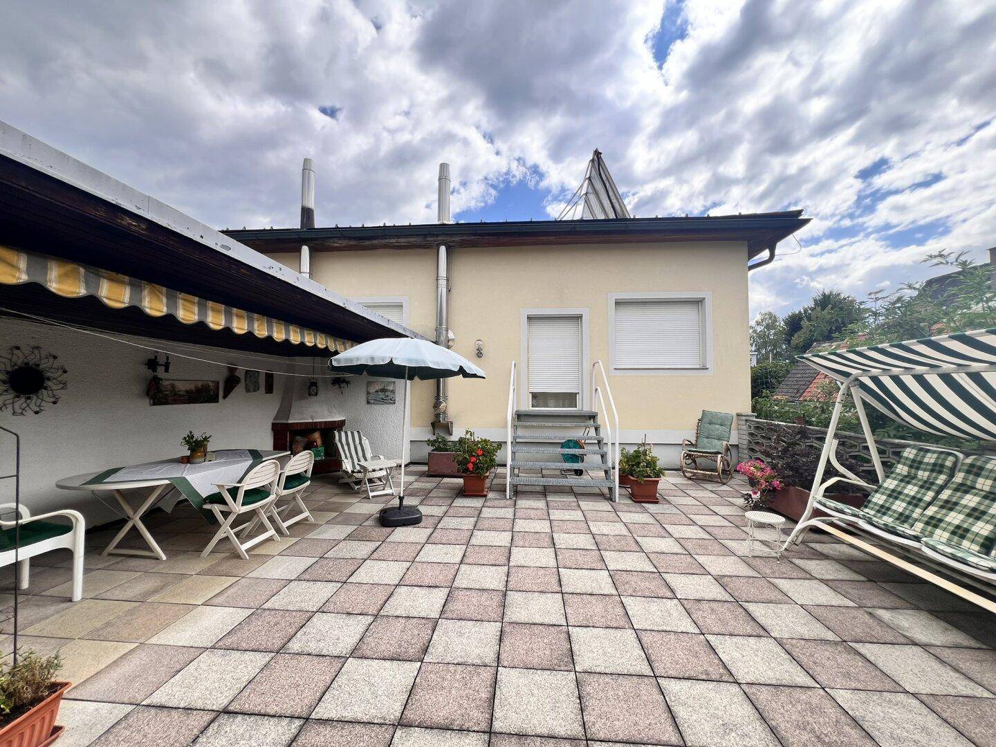 Geräumige geflieste Terrasse mit Gartenmöbeln, Sonnenschirm und Schaukel.