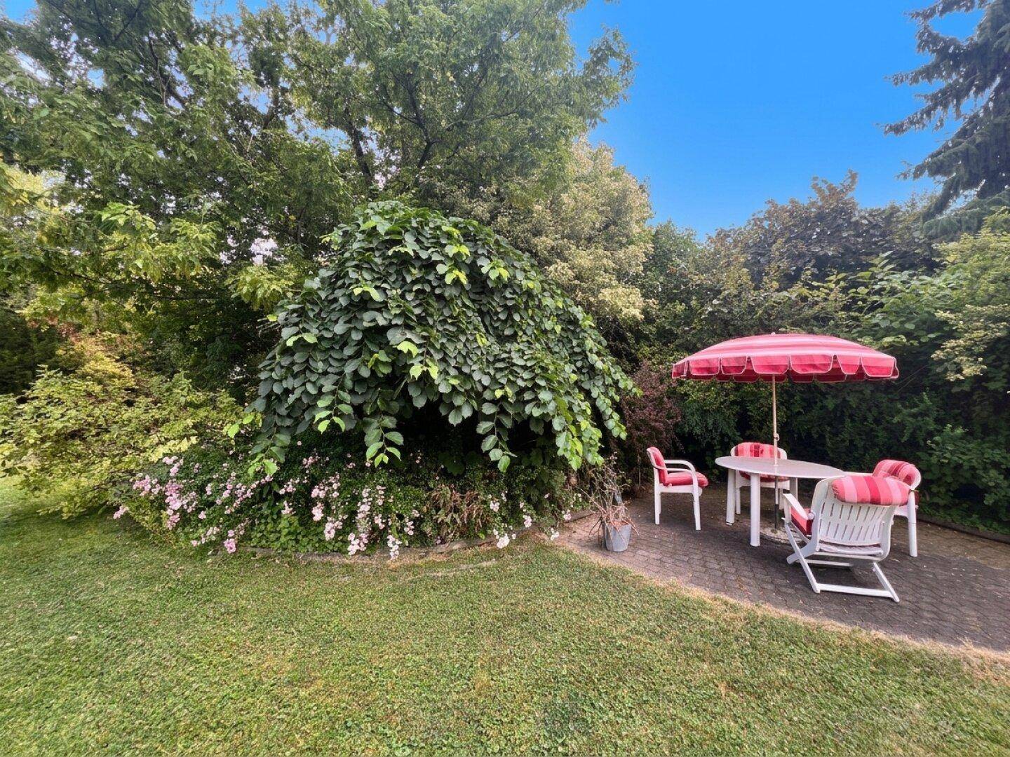 Entspannter Sitzbereich im Garten unter Bäumen mit rotem Sonnenschirm und Gartenmöbeln.