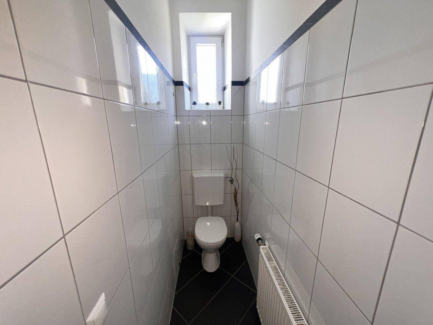 Schmales WC mit weißen Wandfliesen, kleinem Fenster und Heizkörper.