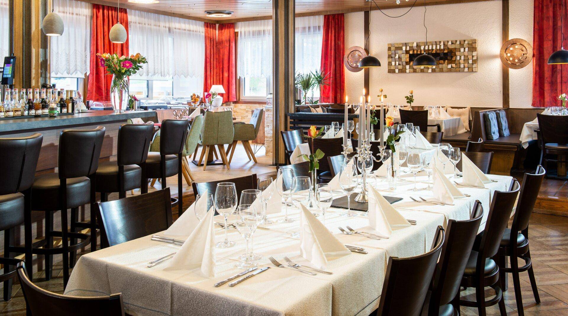 Festlich gedeckter langer Tisch in einem eleganten Speisesaal mit Bar im Hintergrund und roten Vorhängen.
