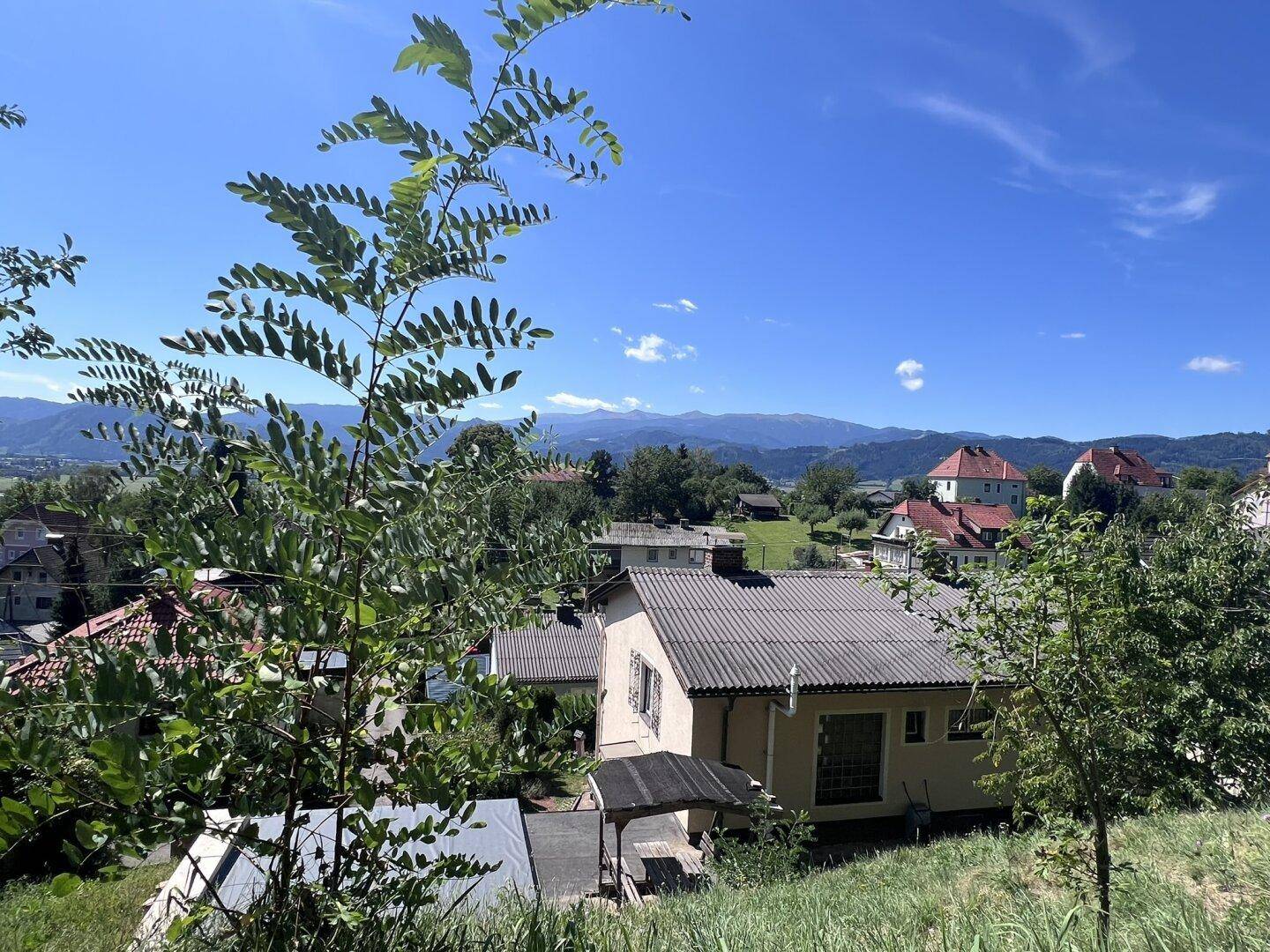 Weitläufiger Blick über die hügelige Landschaft mit Häusern und Bergen im Hintergrund an einem sonnigen Tag.