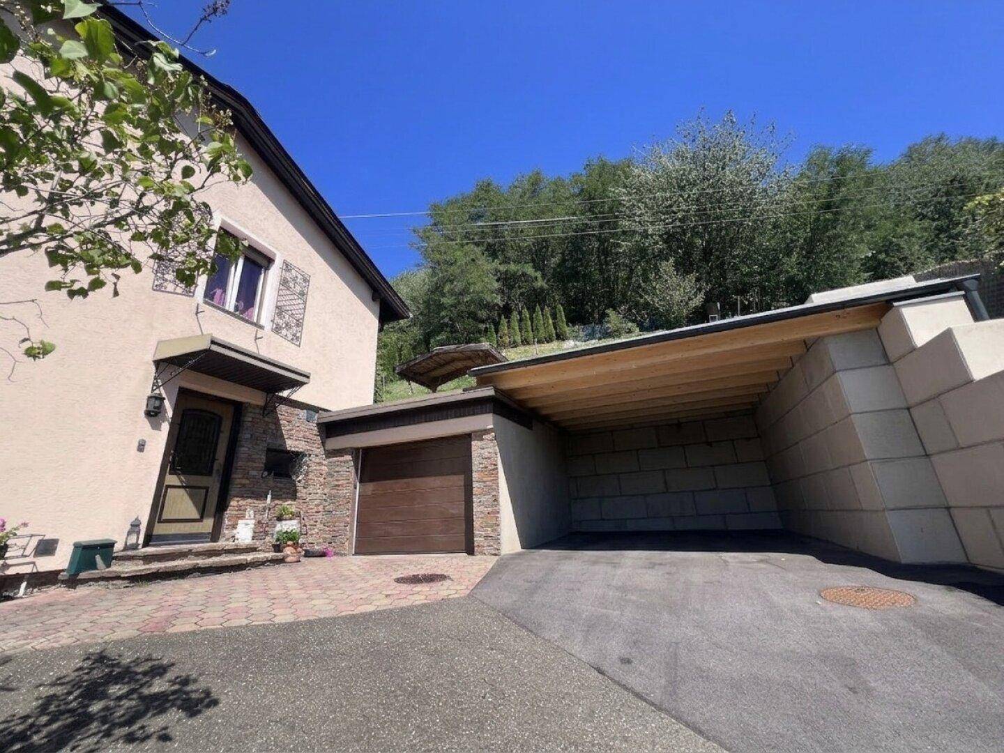 Seitenansicht des Hauses mit Garage, Carport und Stützmauer, umgeben von viel Grün.