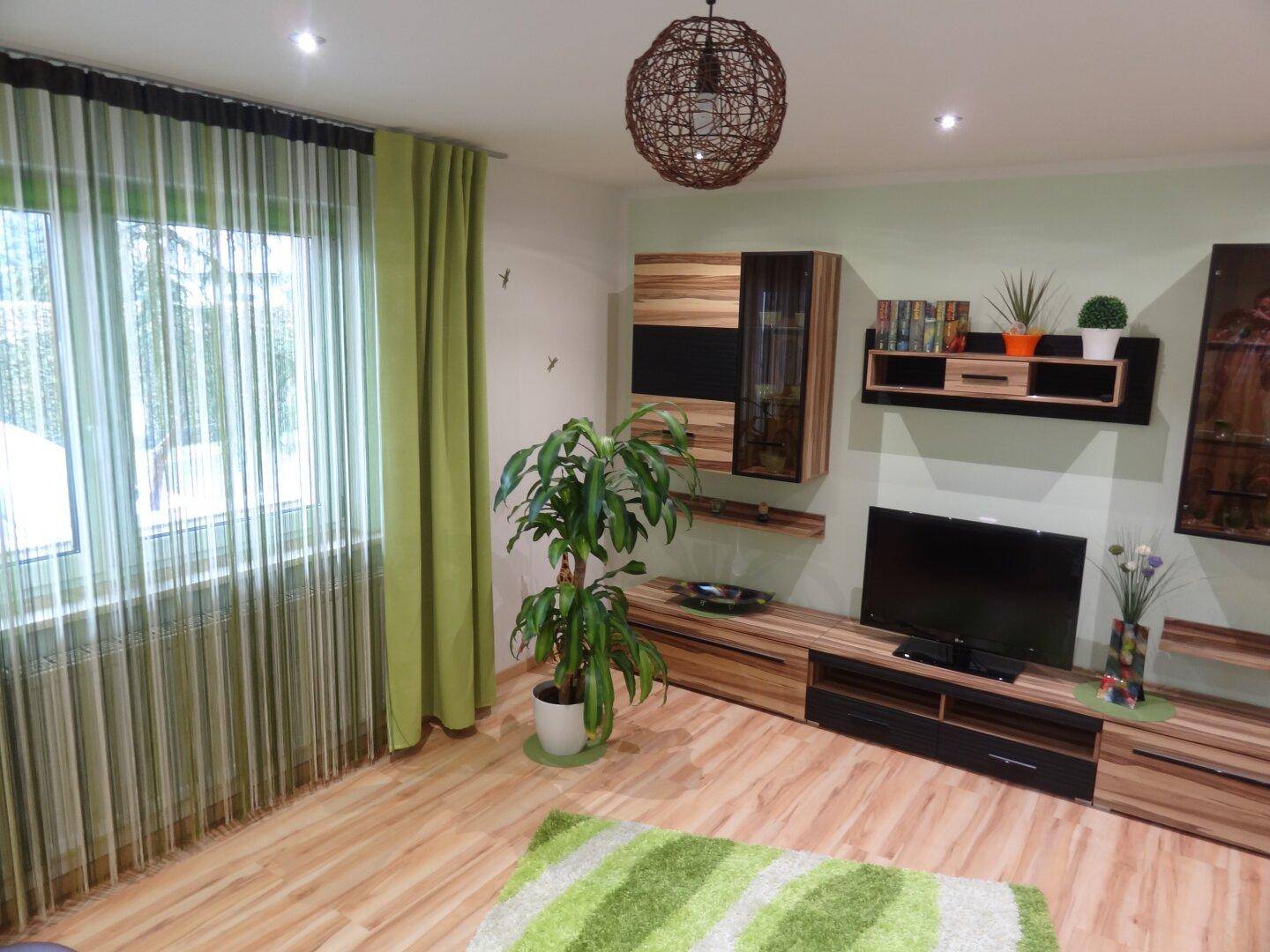 Gemütliches Wohnzimmer mit Holzmöbeln, Fernseher, grünen Vorhängen und Teppich.