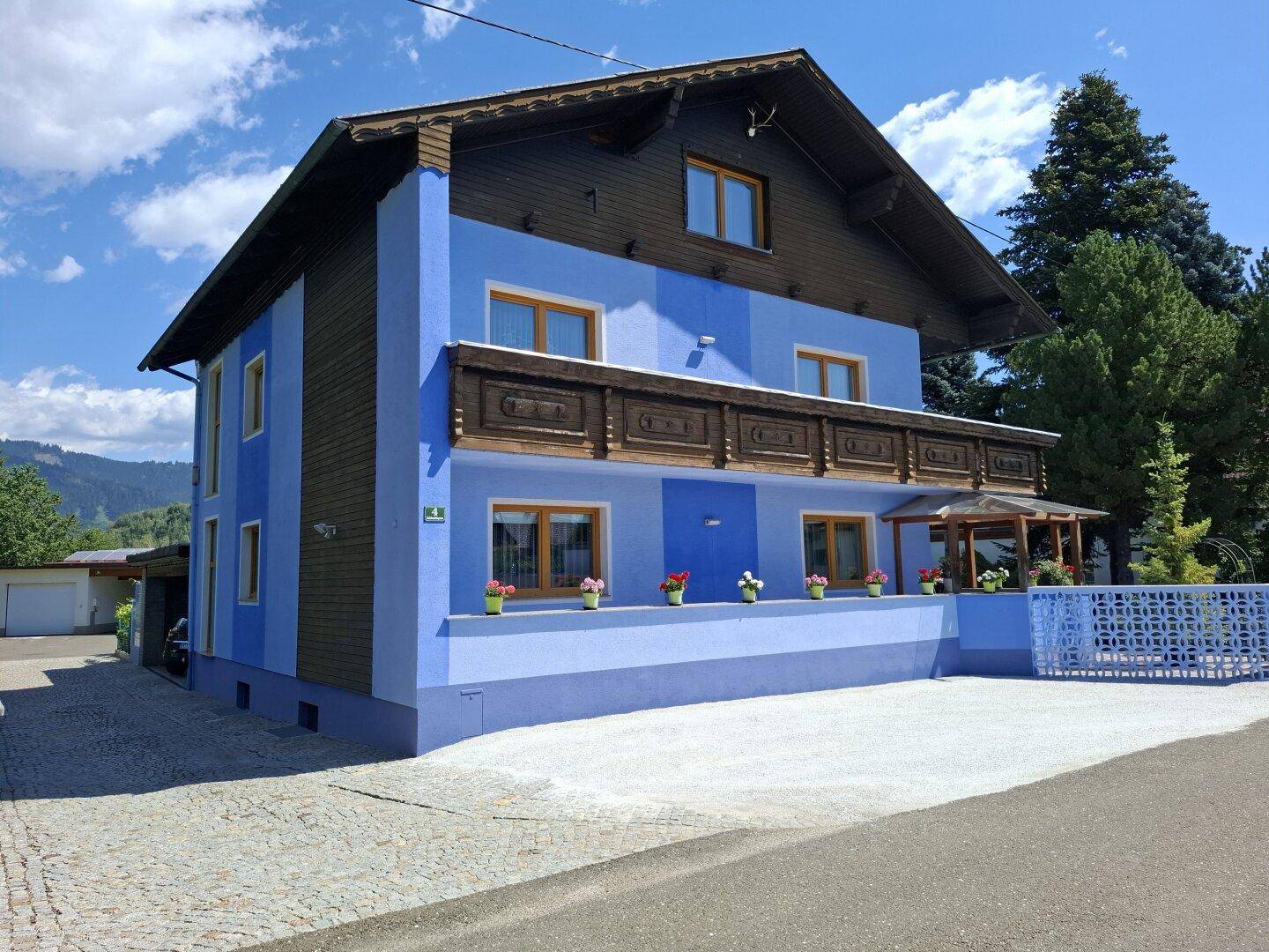 Ansicht des blauen Hauses mit Holzbalkon und gepflasterter Auffahrt unter blauem Himmel.