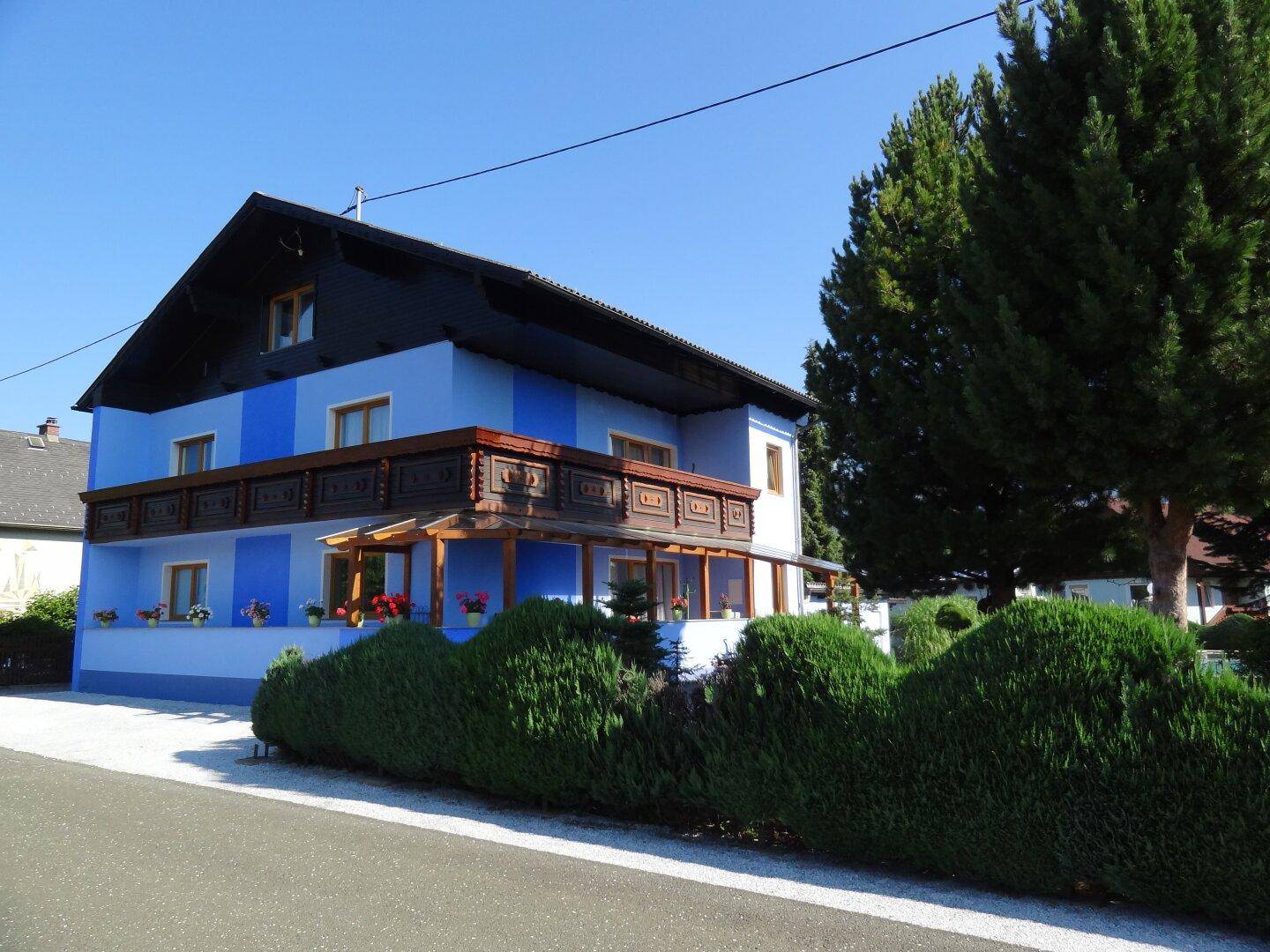 Das blaue Haus von der Straße aus gesehen, mit Holzbalkon und grüner Hecke im Vordergrund.