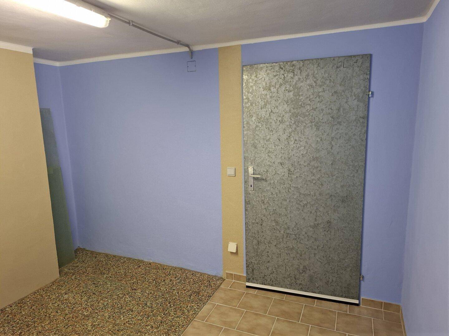 Abstellraum mit hellblauen Wänden, einer robusten Metalltür und gemischtem Bodenbelag.