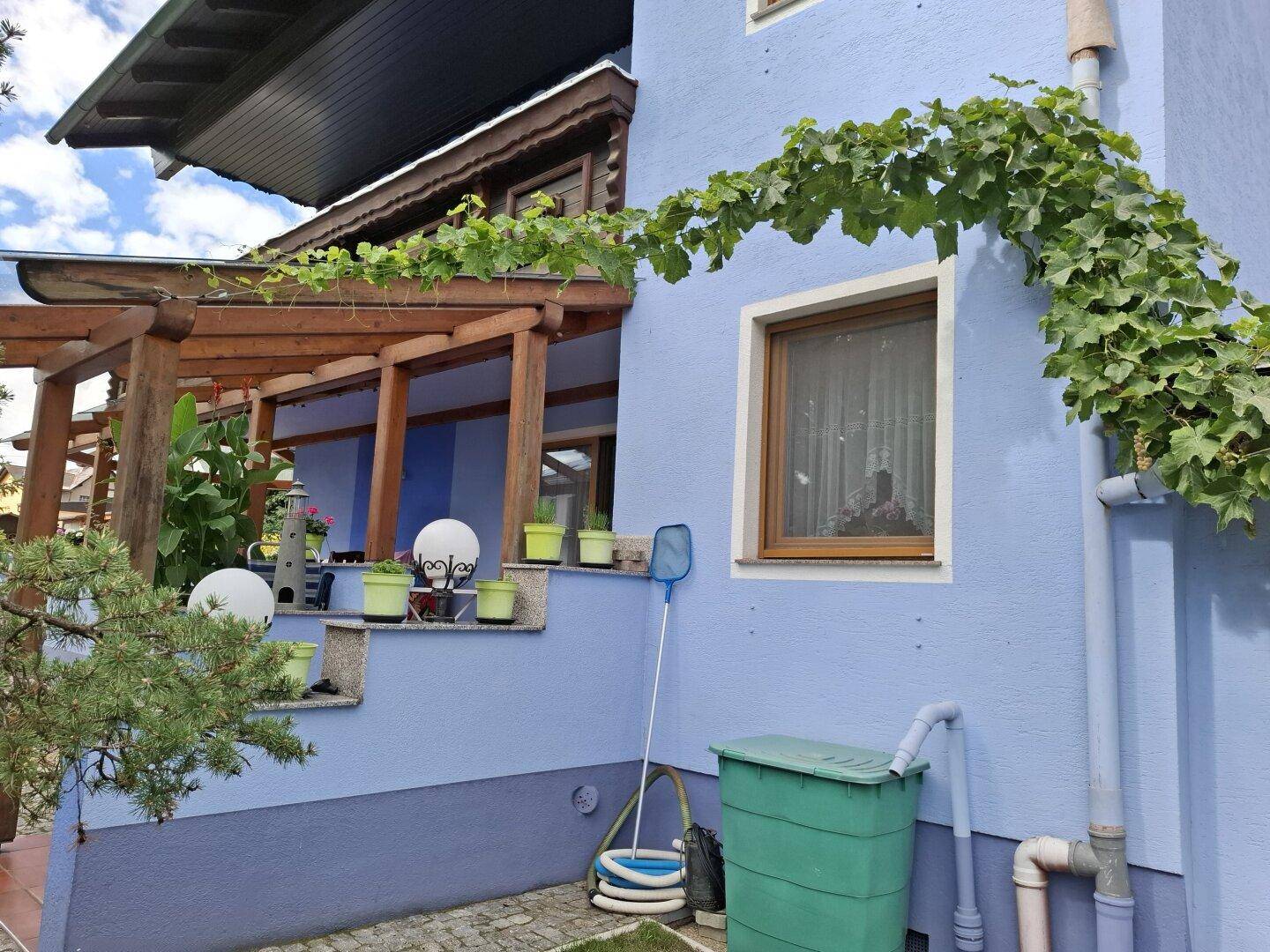 Blaue Hausfassade mit einer überdachten Holzterrasse, begrünt mit Kletterpflanzen und einem Regenfass.