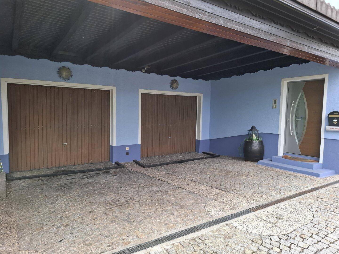 Ansicht der Doppelgarage und des Haupteingangs mit gepflasterter Auffahrt und blauer Fassade.