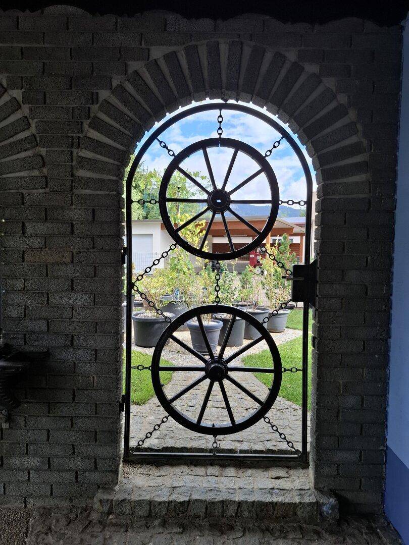 Dekoratives Metalltor mit Wagenradmotiven in einem Ziegelbogen, das zum Garten führt.
