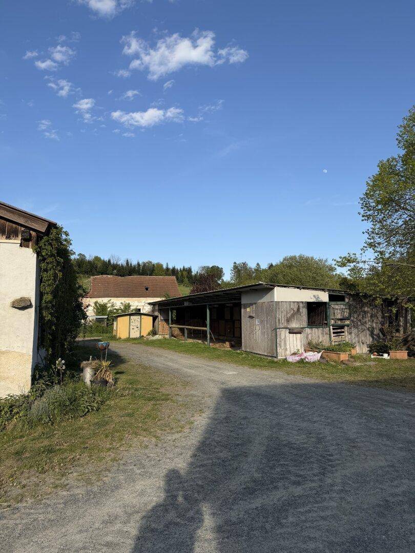 Ein ländlicher Hof mit mehreren rustikalen Gebäuden und einem Kiesweg unter einem teilweise bewölkten Himmel.