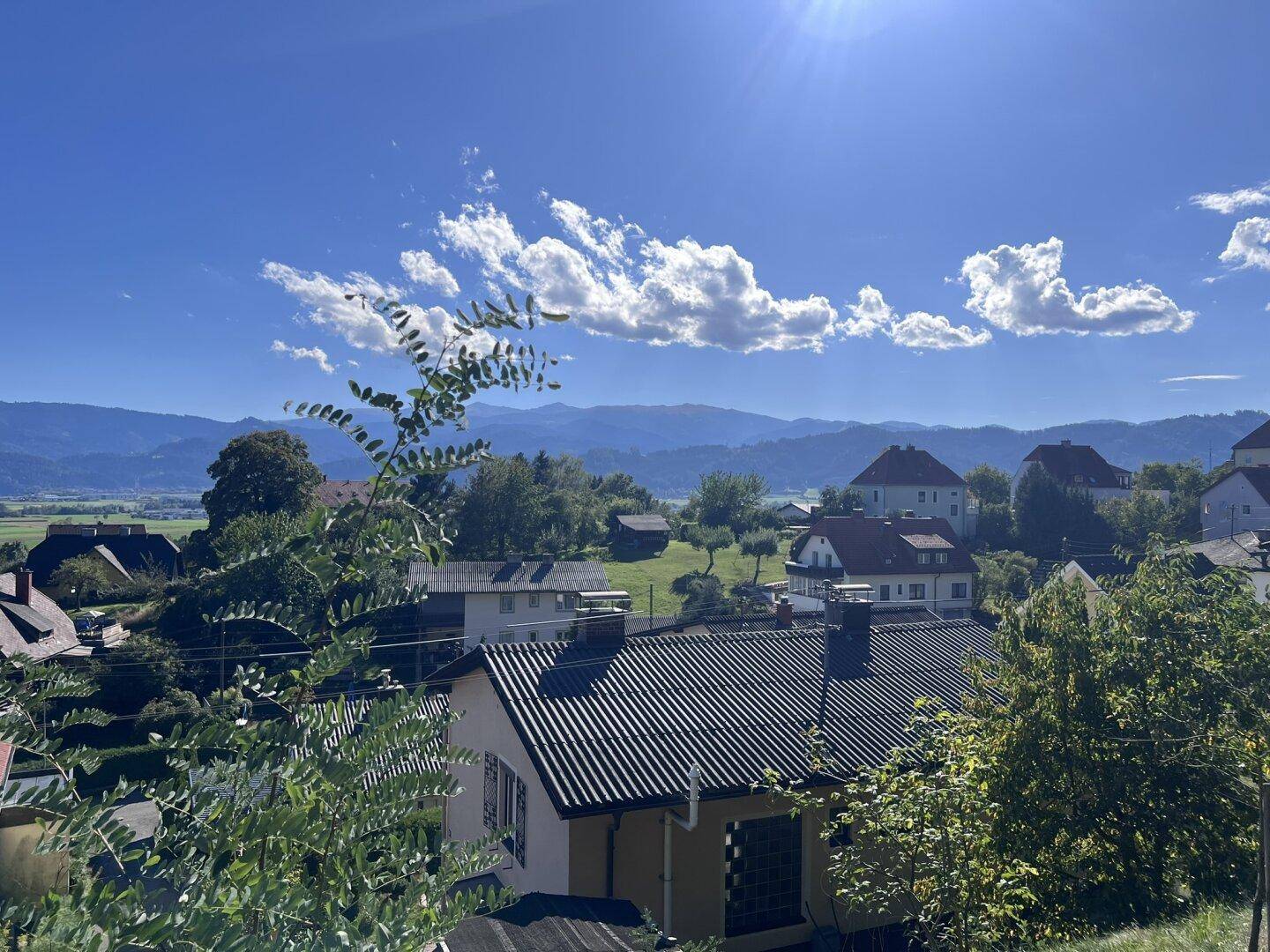 Weitläufiger Blick über eine ländliche Siedlung mit Bergen im Hintergrund unter strahlend blauem Himmel.