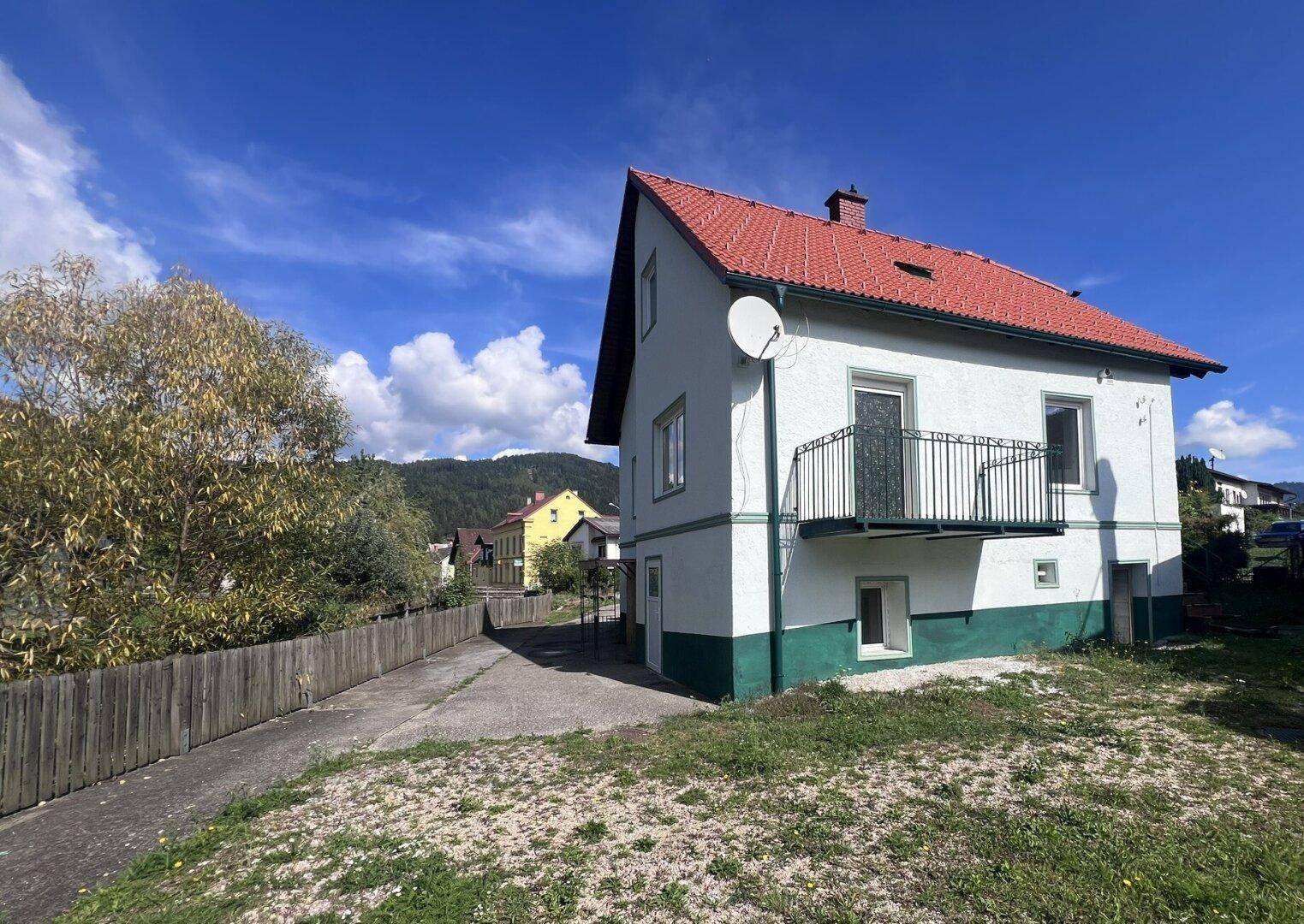 Ansicht des Hauses mit rotem Ziegeldach, weißer Fassade und Balkon vor bewaldeter Hügellandschaft.