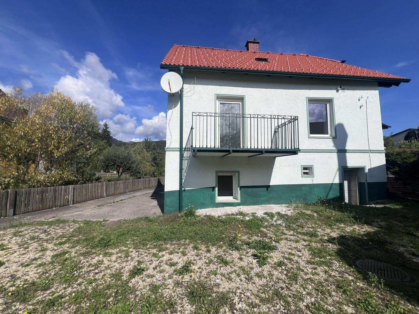 Freistehendes Haus mit rotem Dach, weißer Fassade und grünem Sockel in ländlicher Umgebung.
