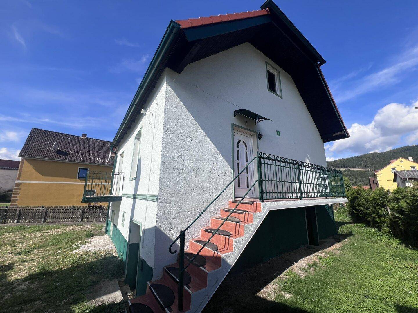 Vorderansicht des Hauses mit Außentreppe, Balkon und Eingangstür unter einem Vordach.