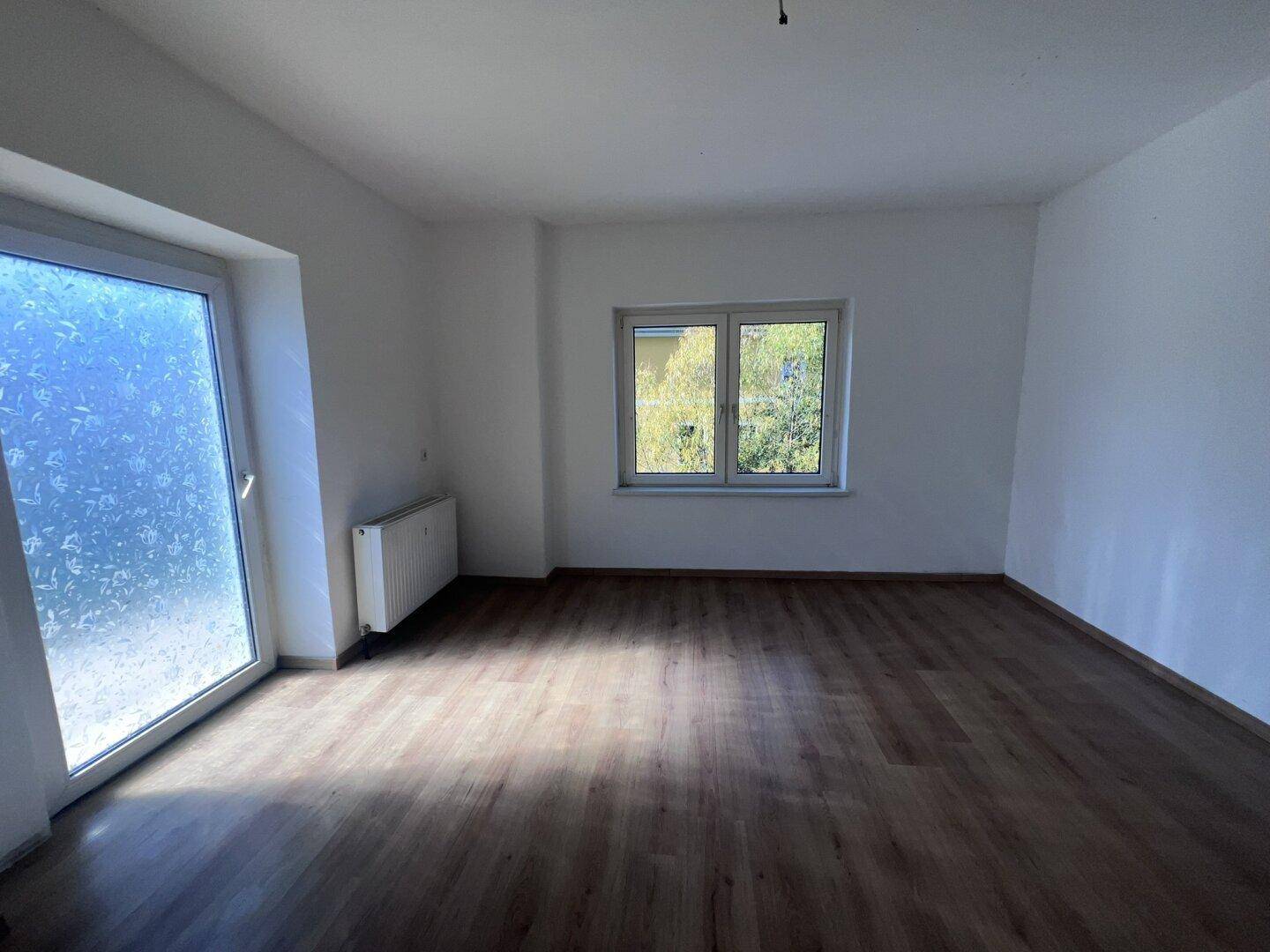 Geräumiges, leeres Zimmer mit Laminatboden, weißen Wänden und viel Tageslicht durch Fenster.