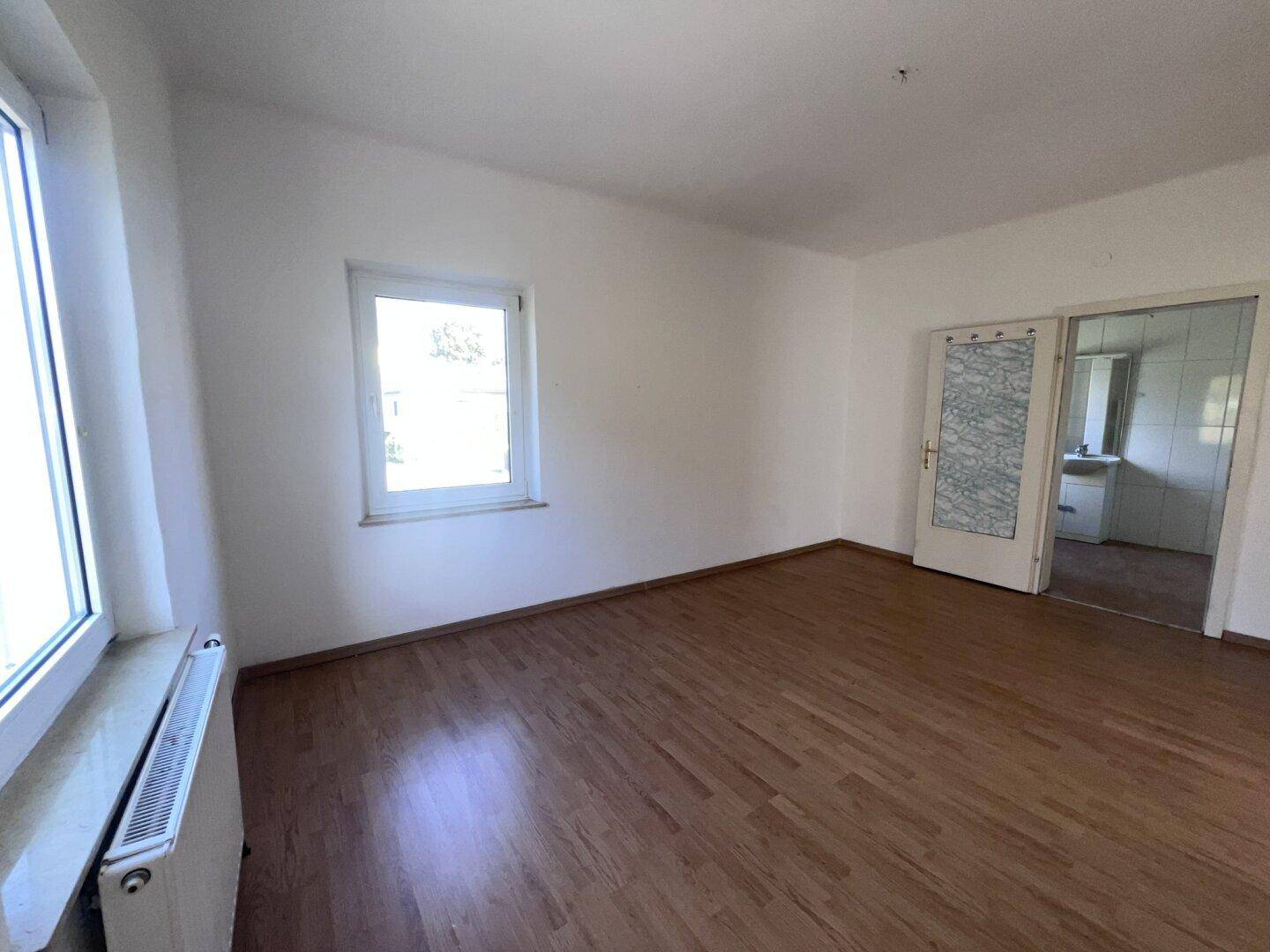 Leeres Zimmer mit Laminatboden, weiß gestrichenen Wänden und Zugang zu einem weiteren Raum.