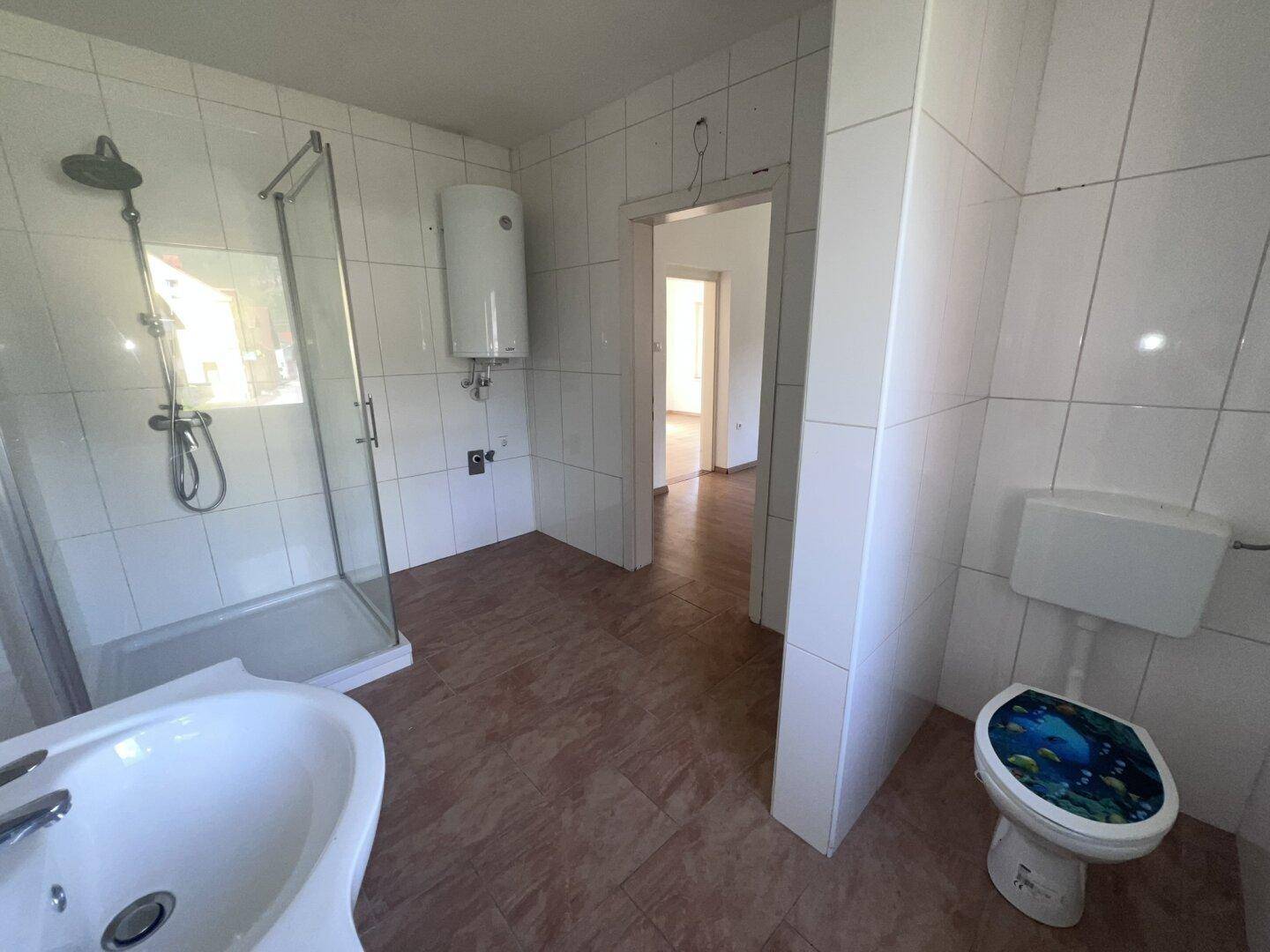 Badezimmer mit Duschkabine, Toilette und Boiler, weiß gefliest und mit braunem Boden.