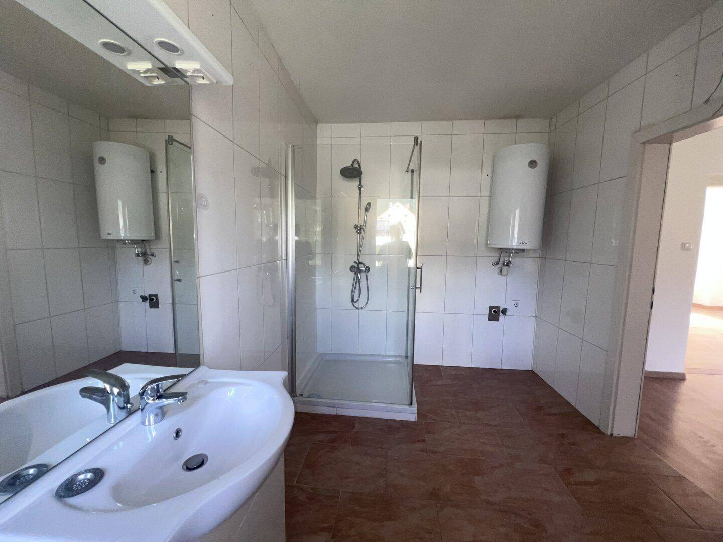 Badezimmer mit Waschbecken, Spiegel, Duschkabine und Boiler, hell gefliest.