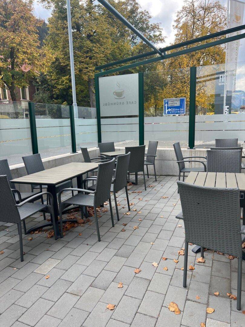 Geräumige Außenterrasse mit zahlreichen Tischen und Stühlen, umgeben von Bäumen und einer Glasabtrennung.