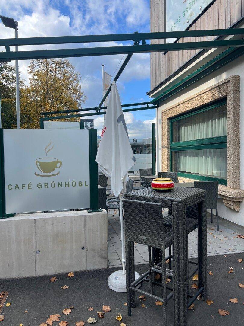 Außenterrasse des Cafés mit Rattanmöbeln, Sonnenschirm und einem Schild mit dem Café-Logo.