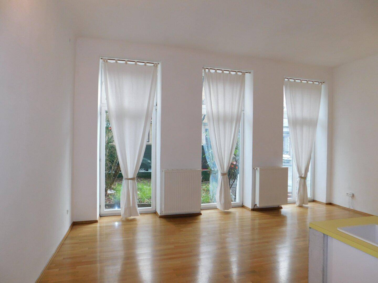 Lichtdurchfluteter Wohnraum mit glänzendem Holzboden und Blick ins Grüne durch hohe Fenster.