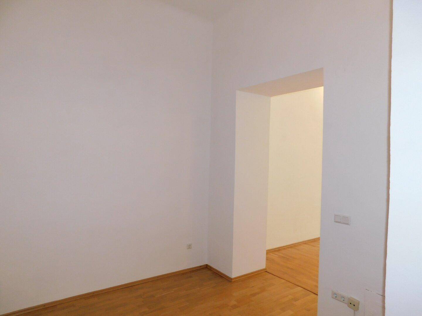 Leerer Raum mit weiß gestrichenen Wänden und hellem Parkettboden, offen zu einem weiteren Bereich.