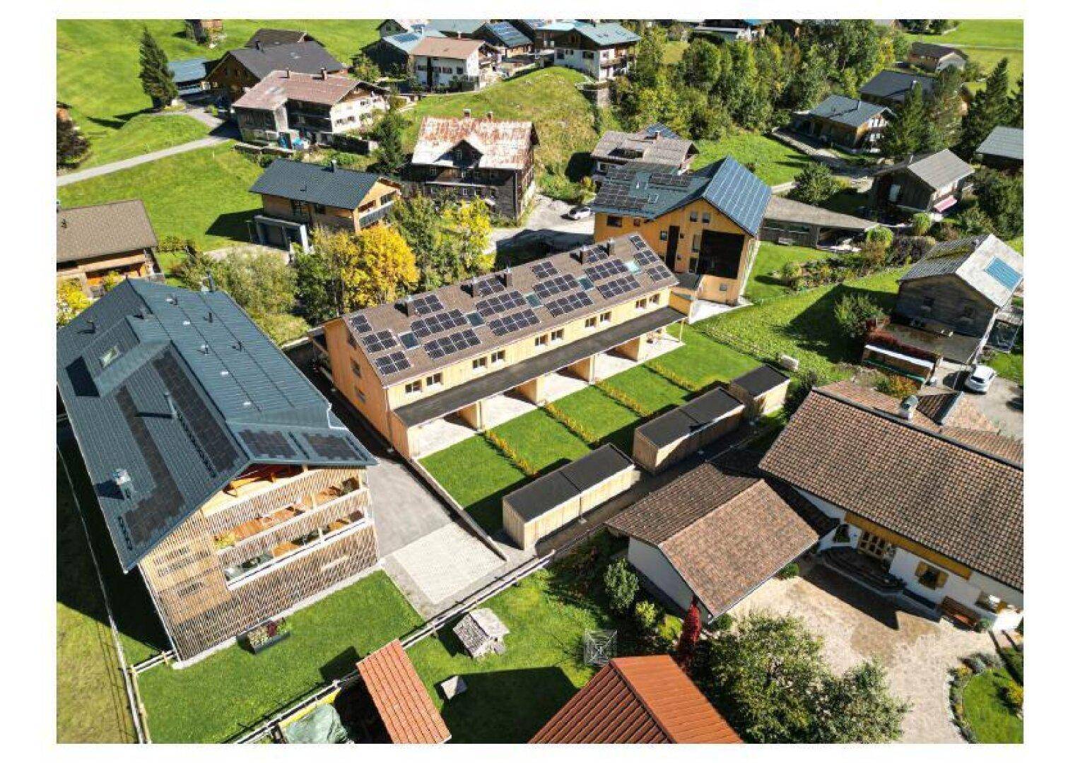 Detaillierte Luftaufnahme der neuen Holzgebäude mit Solaranlagen, eingebettet in die Dorfstruktur.