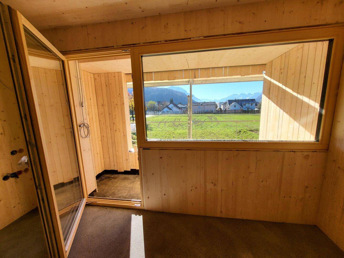 Innenraum im Rohbau mit Holzwänden, großen Fenstern und Blick auf die grüne Landschaft und Berge.