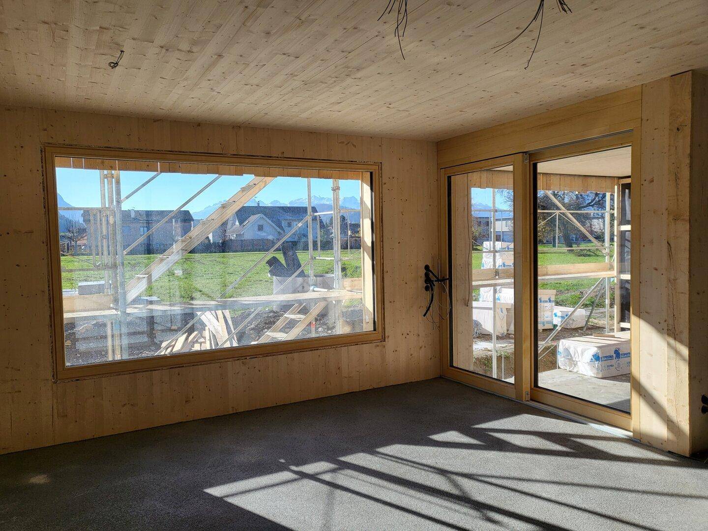 Innenraum im Rohbau mit Holzwänden, großen Fenstern und Schiebetür, die viel Licht hereinlässt.