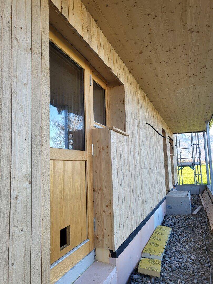 Detailansicht der Holzfassade mit Tür und Fenstern, umgeben von Kies und Baumaterialien.