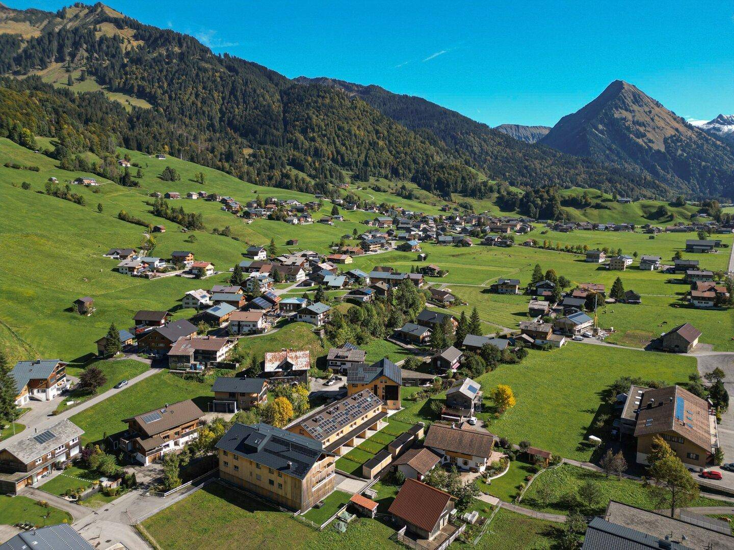 Malerische Luftaufnahme eines Dorfes in einem grünen Tal, umgeben von majestätischen Bergen unter blauem Himmel.