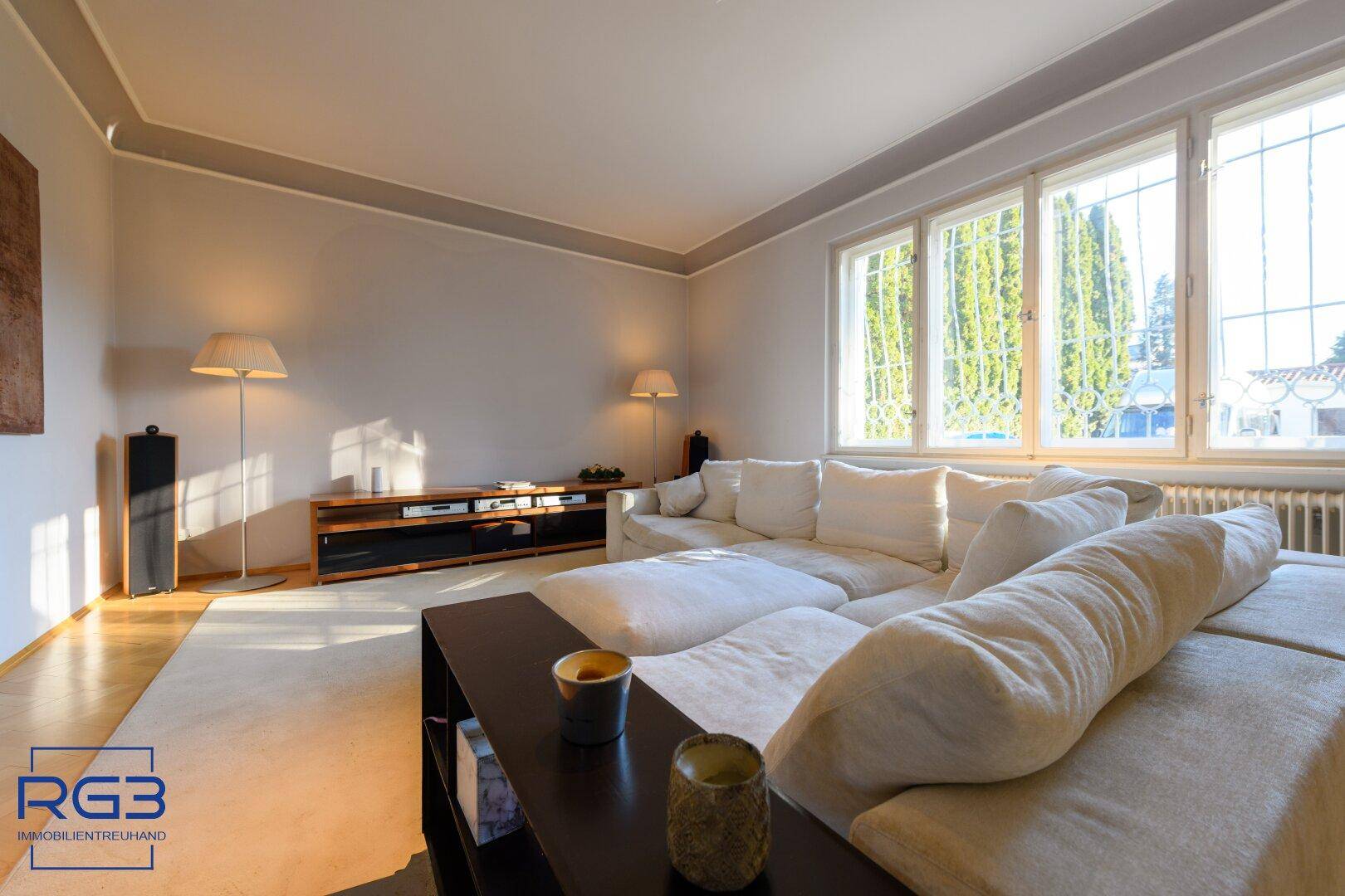 Gemütliches Wohnzimmer mit großem Sofa, Teppichboden und viel Tageslicht durch die Fenster.