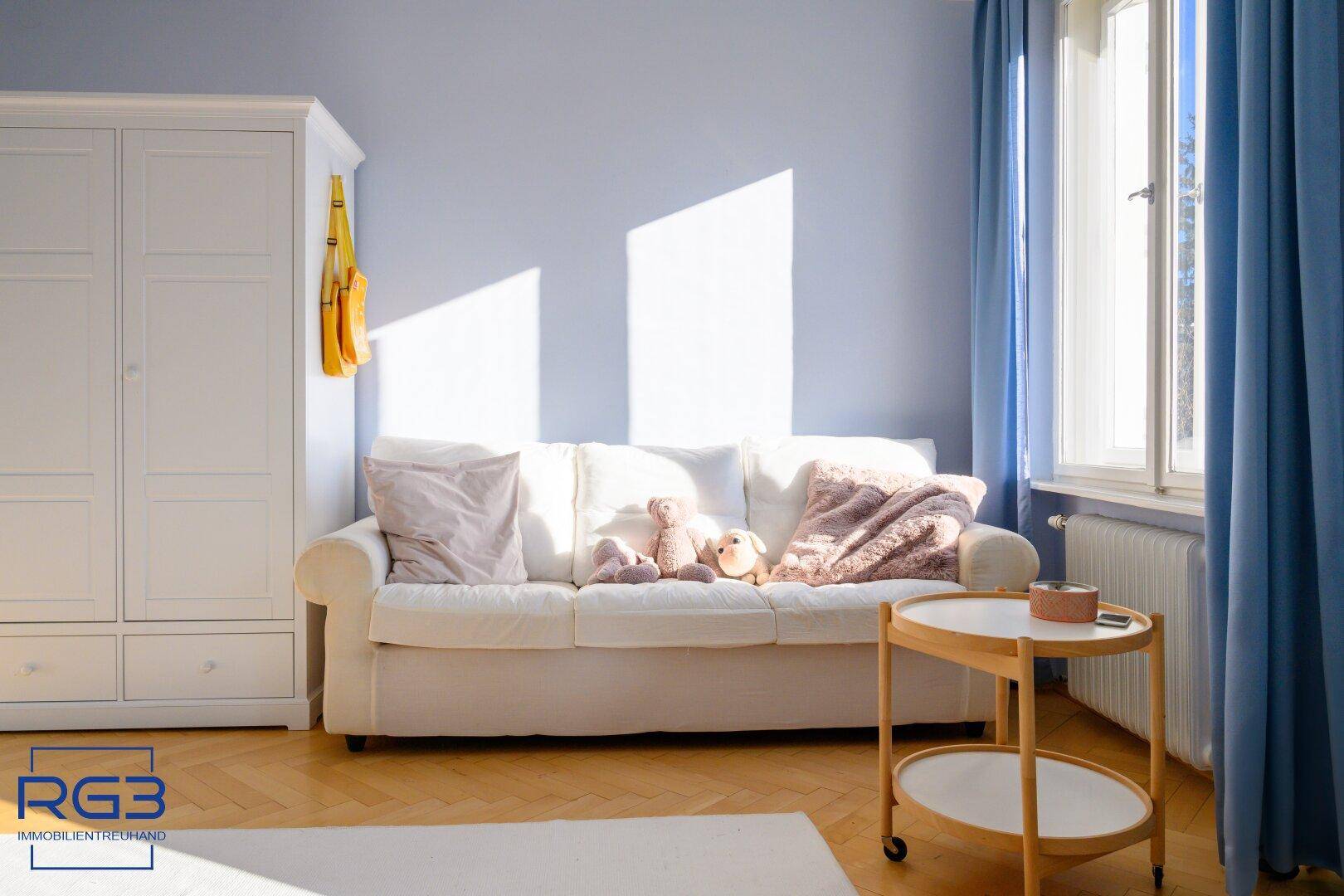 Helles Kinderzimmer mit weißem Schrank, Sofa und blauen Vorhängen.