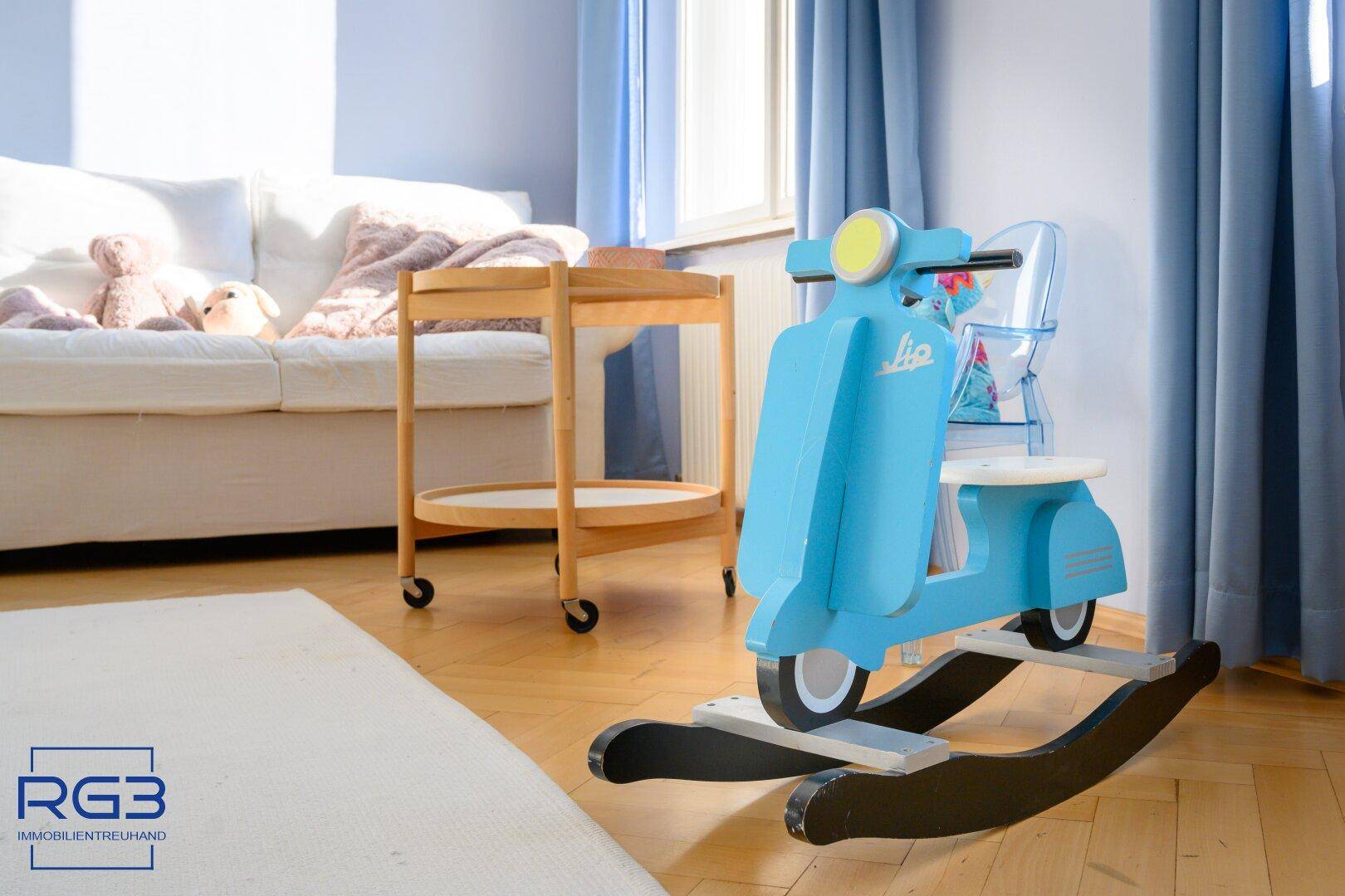 Kinderzimmer mit Holzparkett, weißem Sofa und einem blauen Rollerspielzeug.