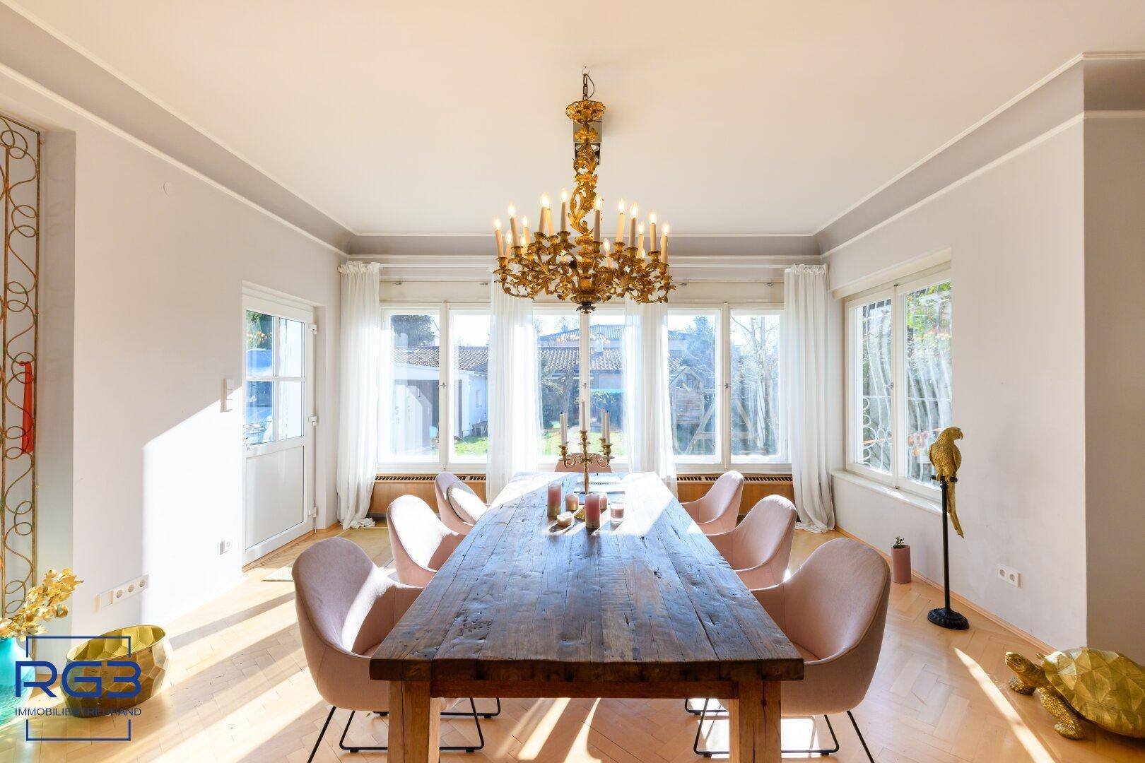 Eleganter Essbereich mit großem Holztisch, Kronleuchter und Blick ins Grüne durch die Fenster.