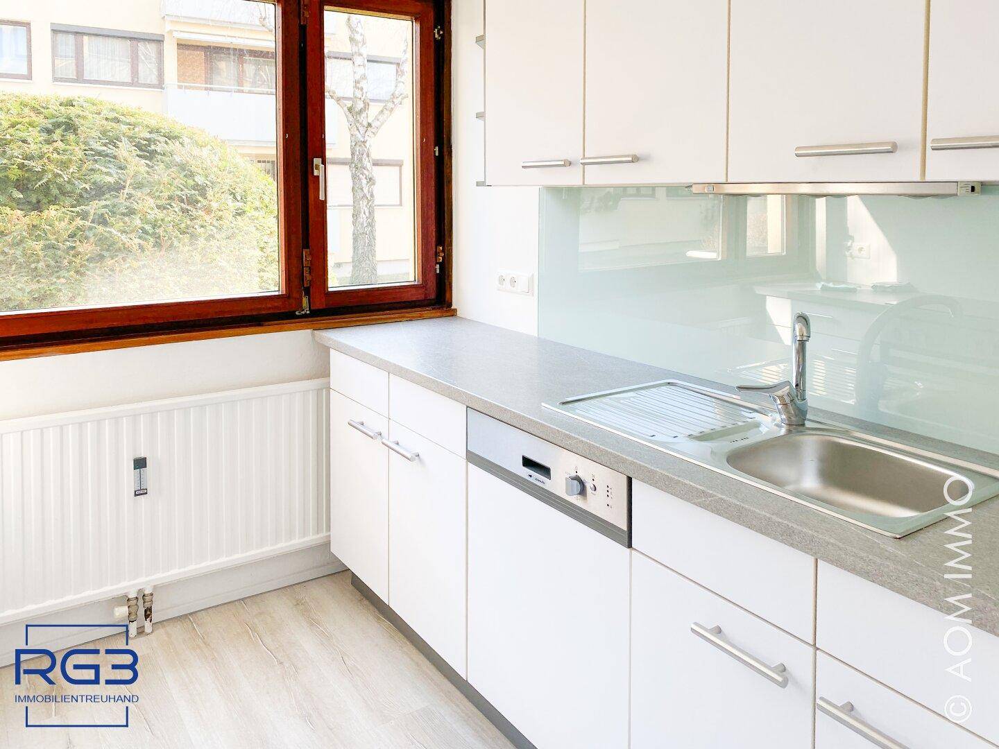 Moderne Küche mit weißen Schränken, Spülmaschine und Fenster mit Blick ins Grüne.