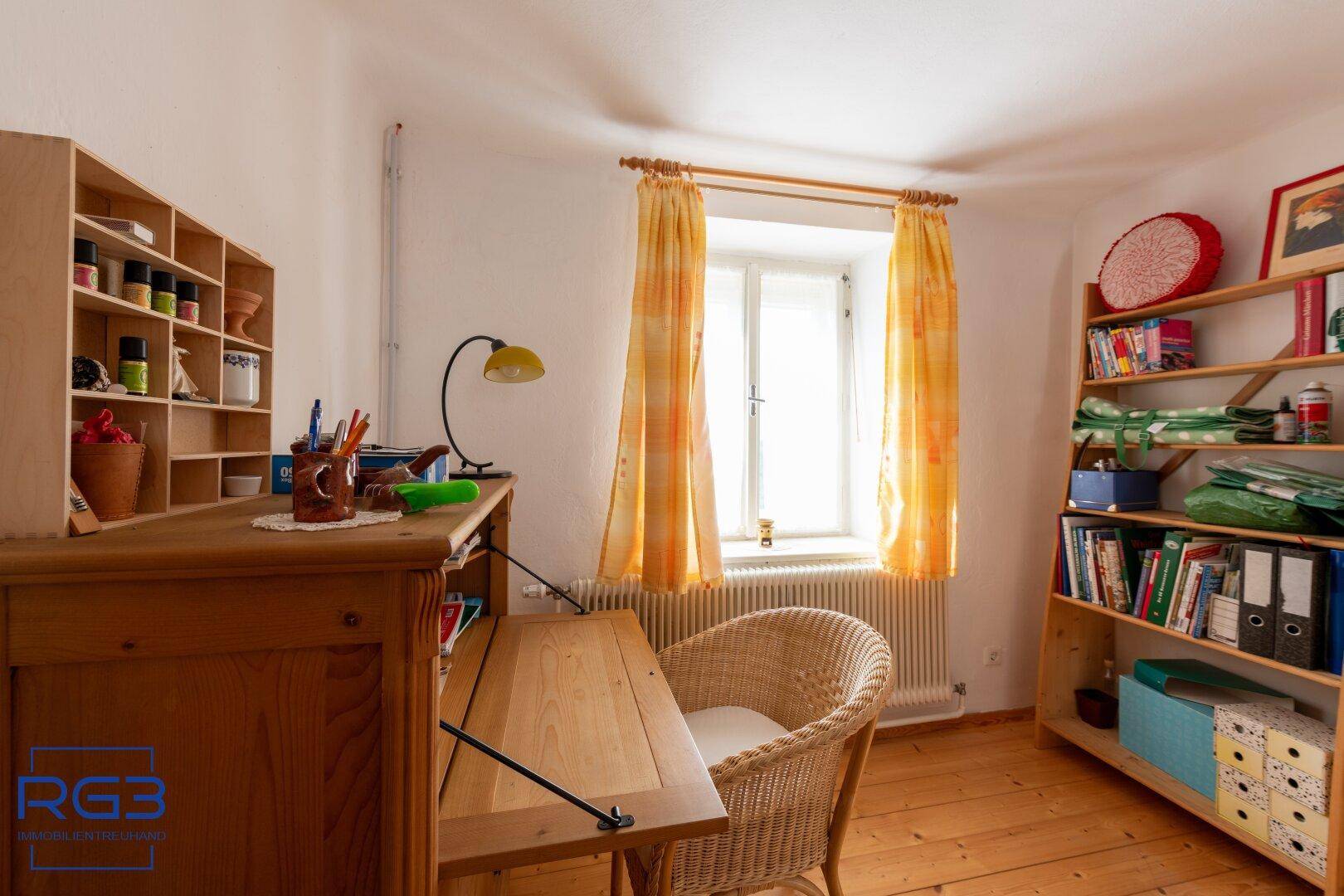 Helles Arbeitszimmer mit Holzmöbeln, Korbstuhl und gelben Vorhängen am Fenster.